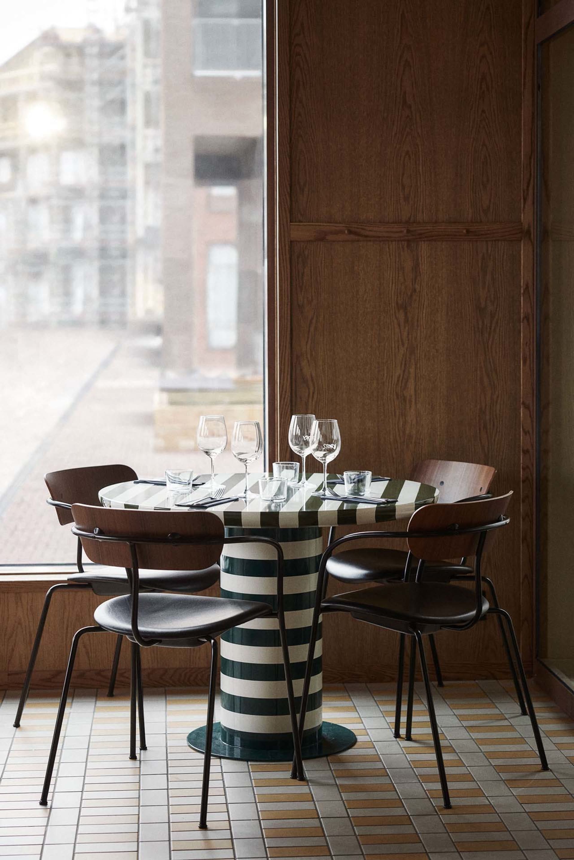 A modern dining setting with a striped table, metal-framed leather chairs, wooden walls, and large windows.