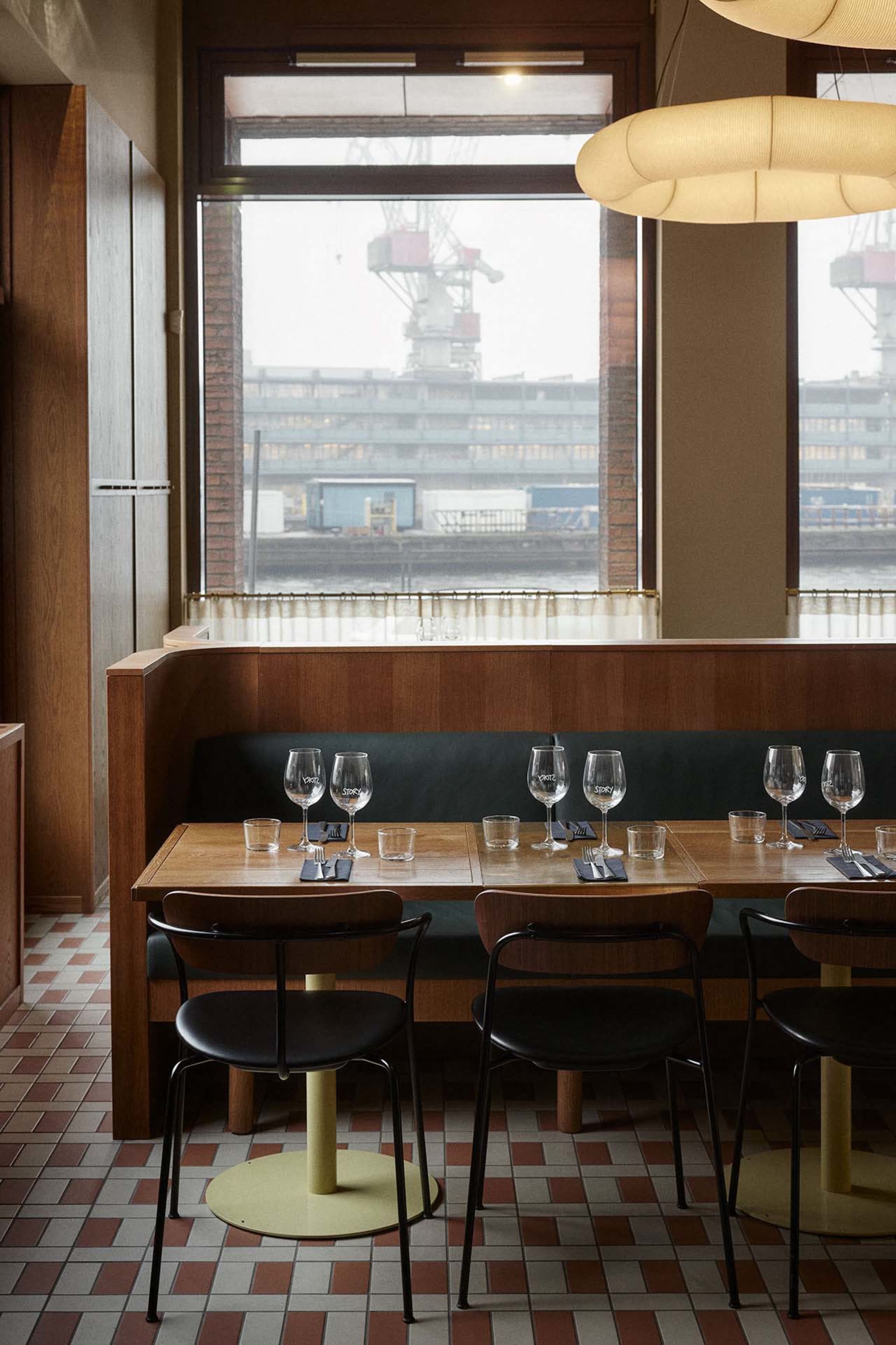 A modern restaurant interior with wooden tables, leather chairs, and large windows overlooking a harbor.