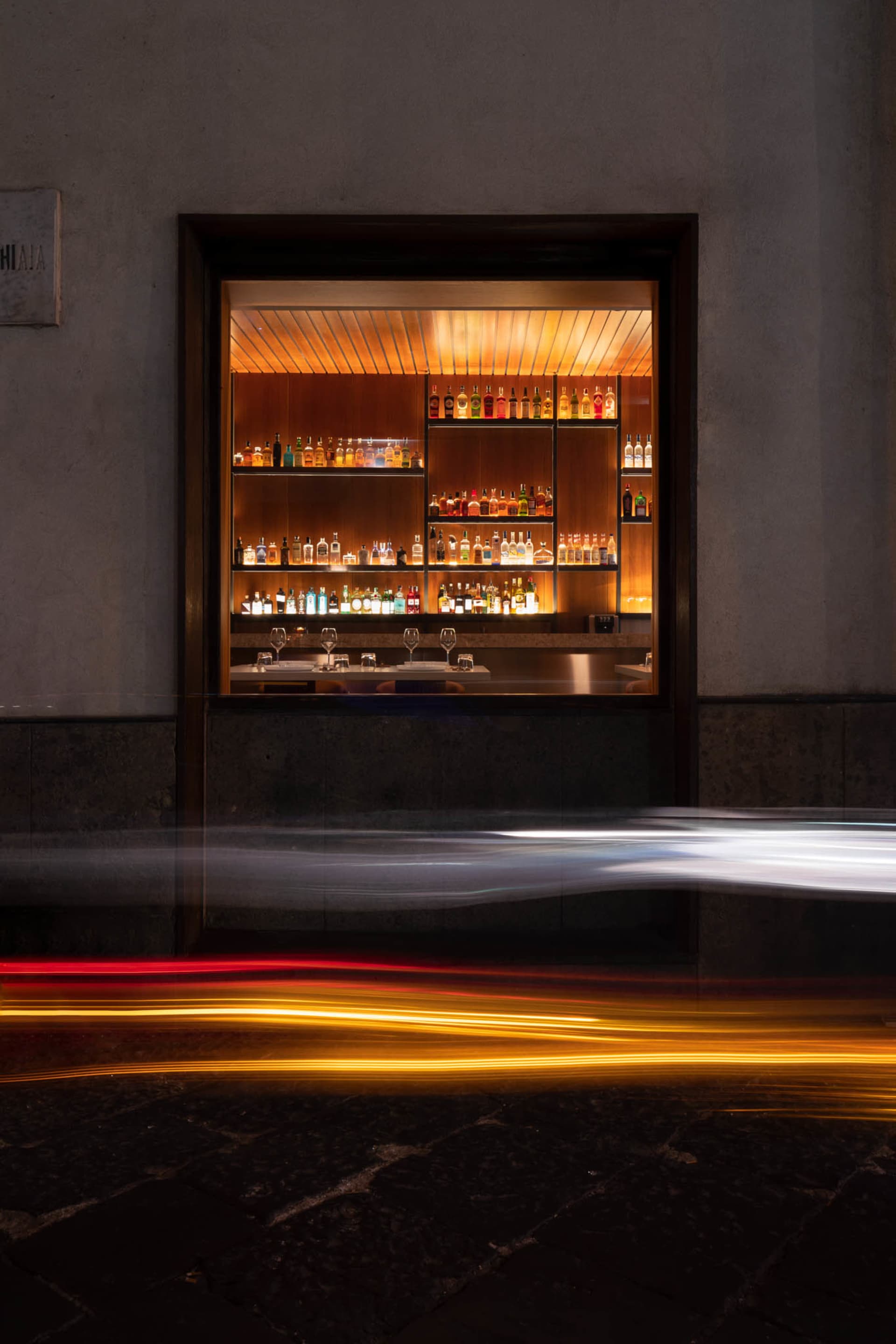 An illuminated restaurant bar with shelves filled with bottles and glasses, set against a dark exterior wall with blurred lig