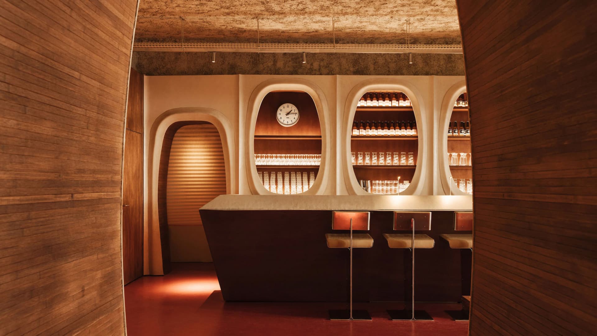 A modern bar interior with a wooden counter, arched walls, and illuminated shelves.