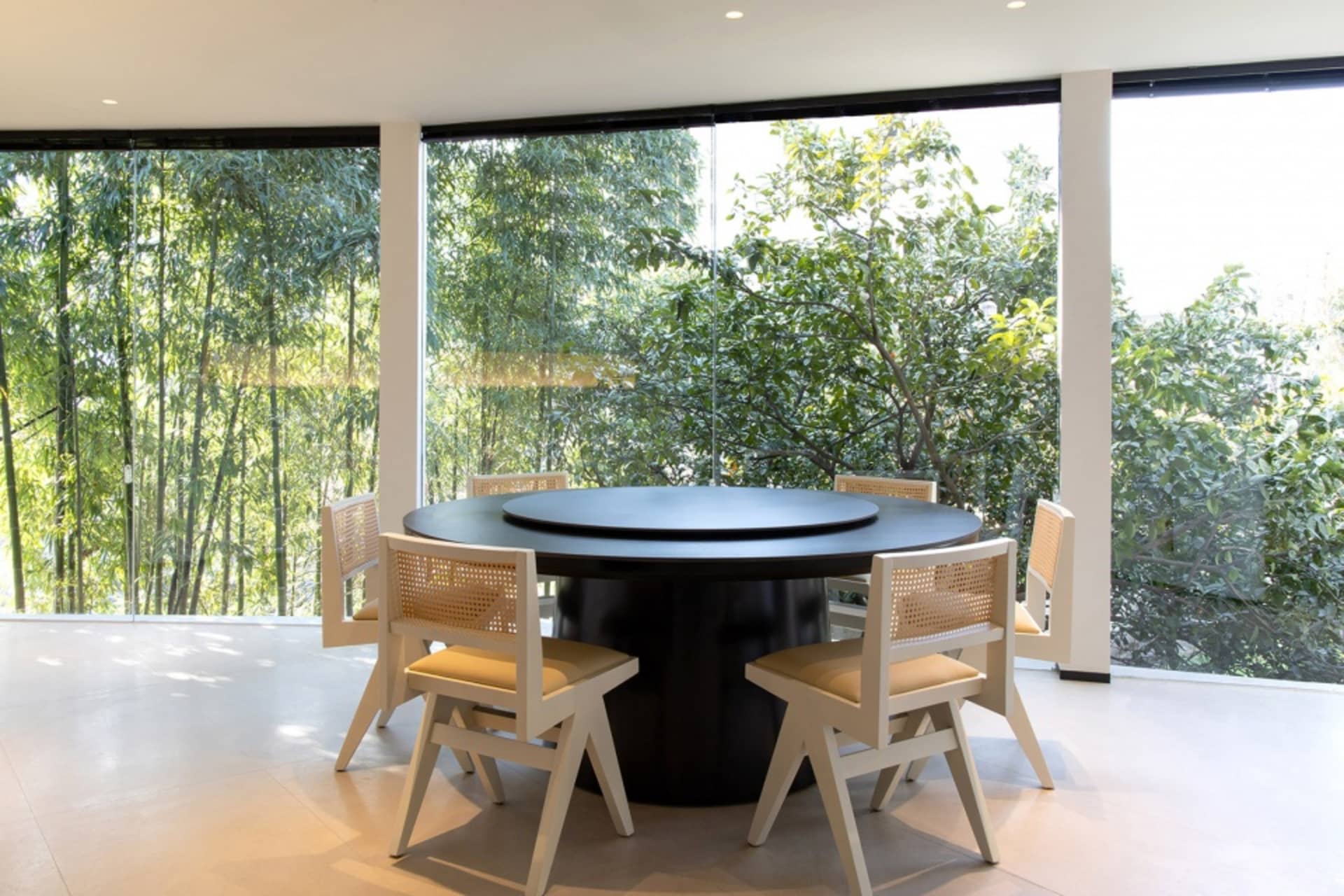 A modern dining room featuring a round table with a glass top and wooden chairs, surrounded by large windows with a view of g