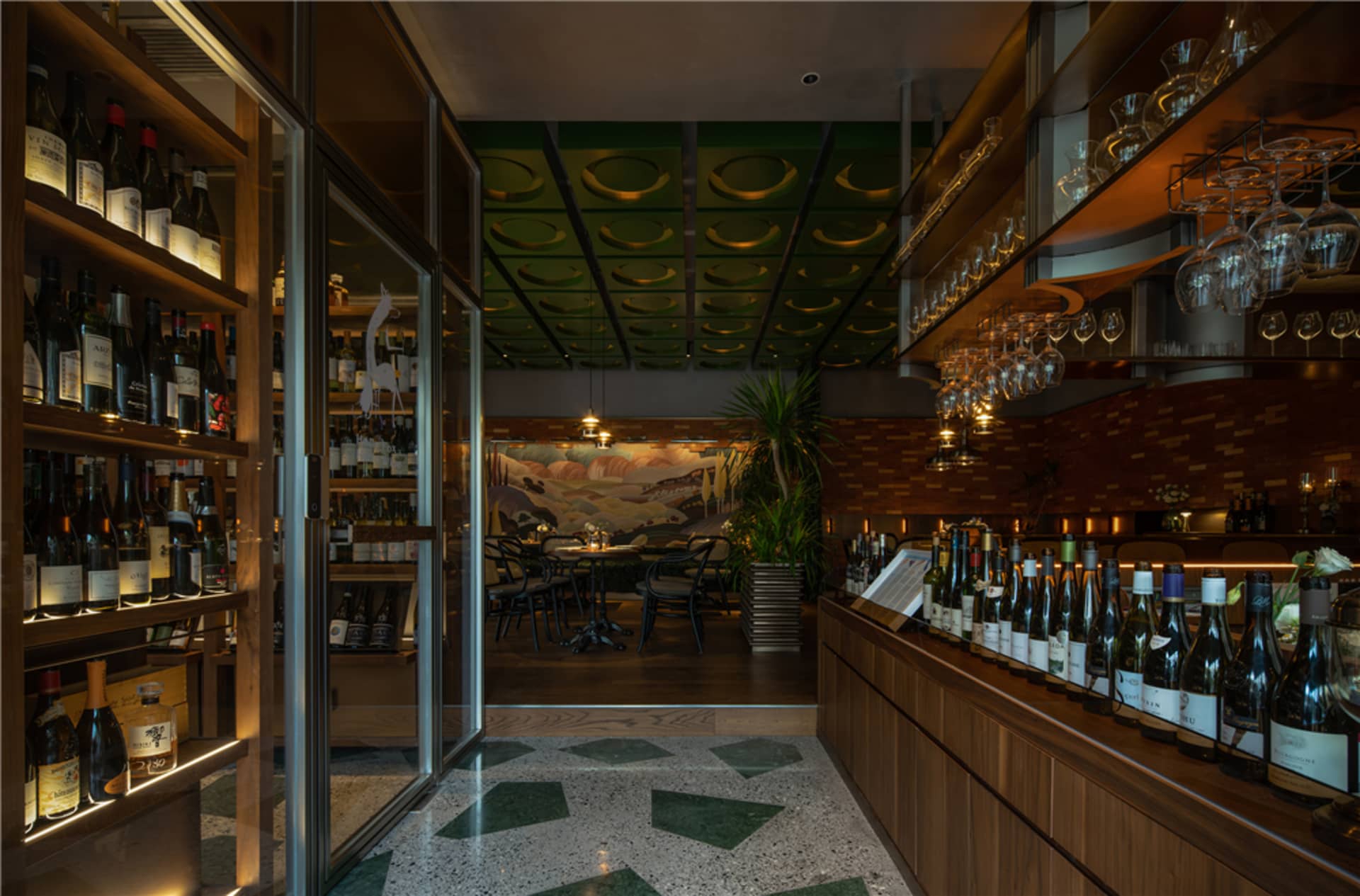Modern wine bar interior with a glass-fronted shelf displaying wine bottles, a bar counter adorned with numerous wine bottles