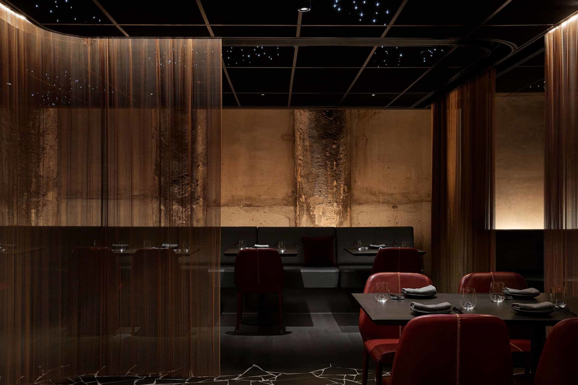 A modern restaurant interior with metal acoustic curtains, dark marble flooring, and dark furniture.