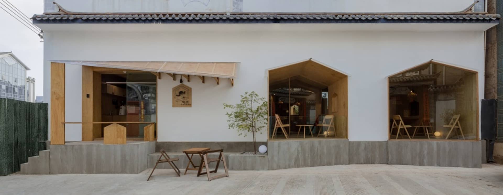 A minimalist façade with wooden accents and glass panels of a restaurant with a concrete base and a tree in front of it.