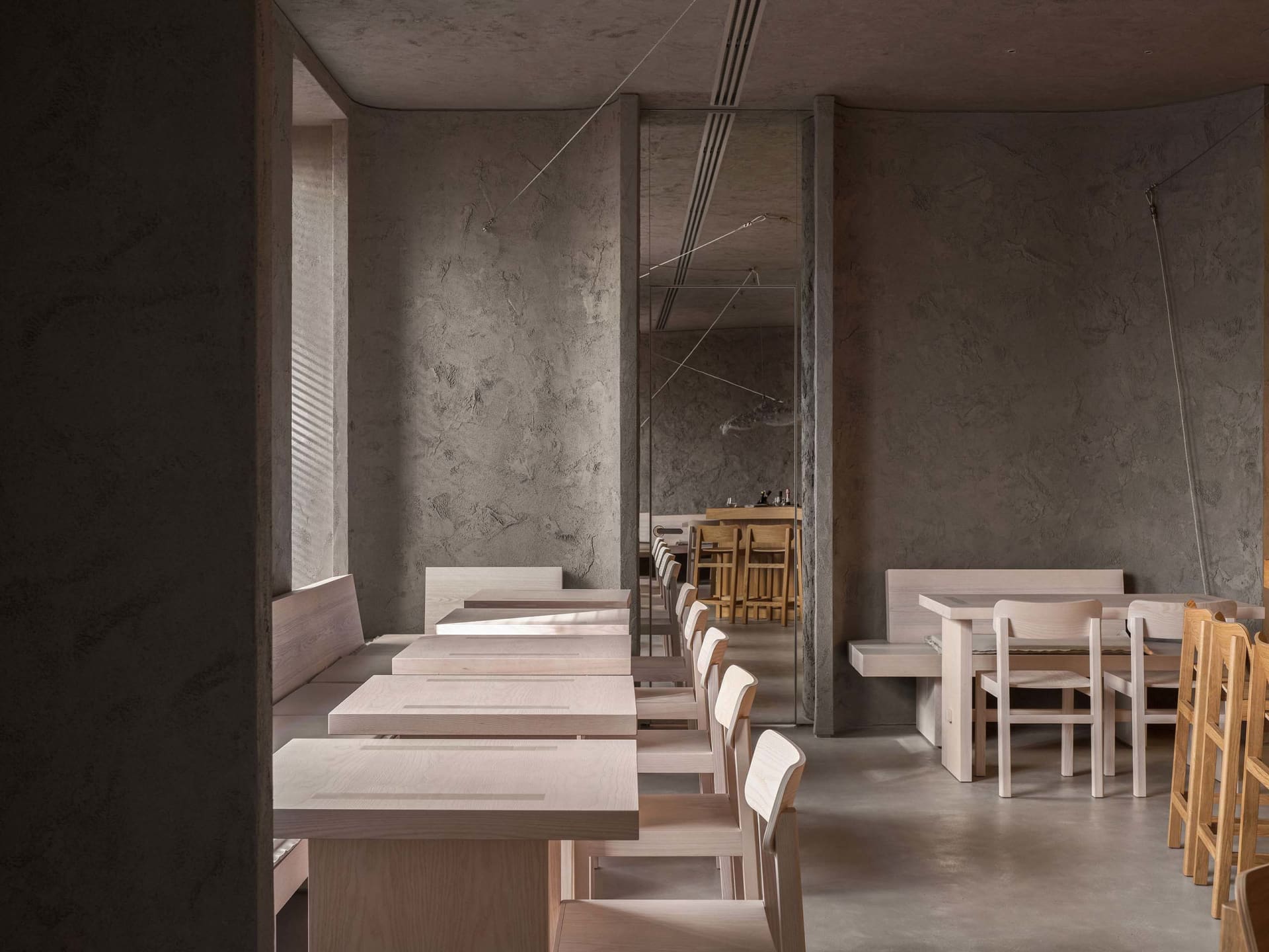 A modern restaurant with wooden tables and benches, concrete walls, and large windows for natural light.