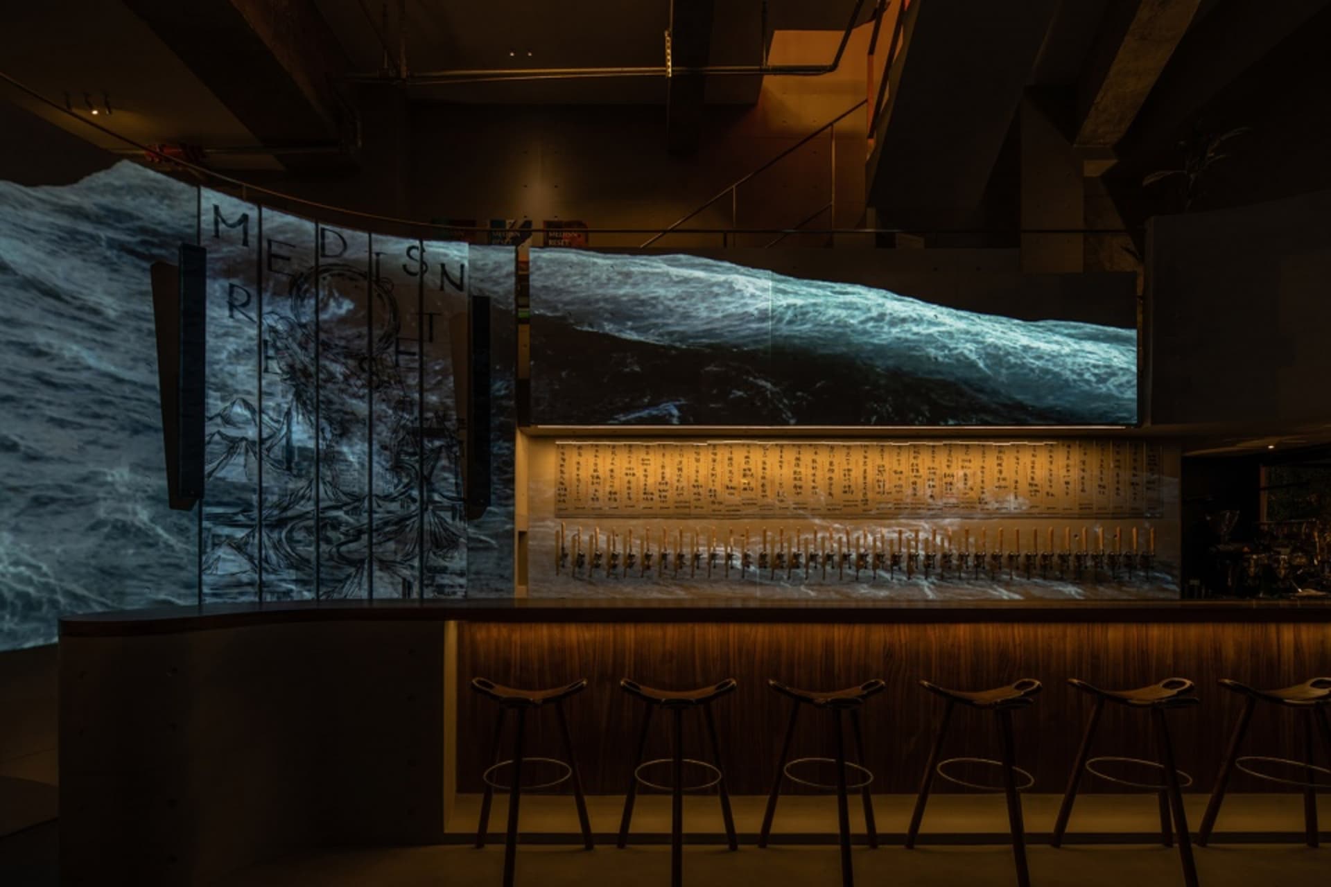 A dimly lit industrial bar with wooden stools and a large screen displaying ocean waves.