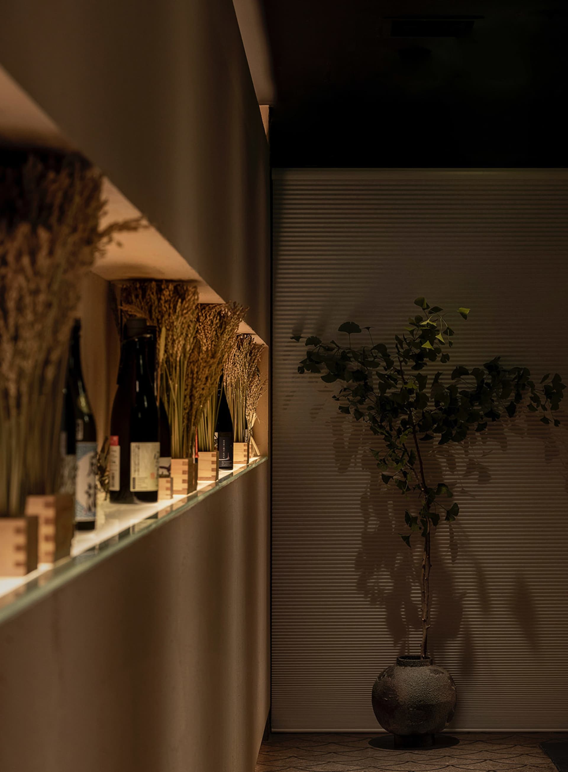Modern minimalist restaurant interior featuring wooden accents, shelf with bottles, and green plant.