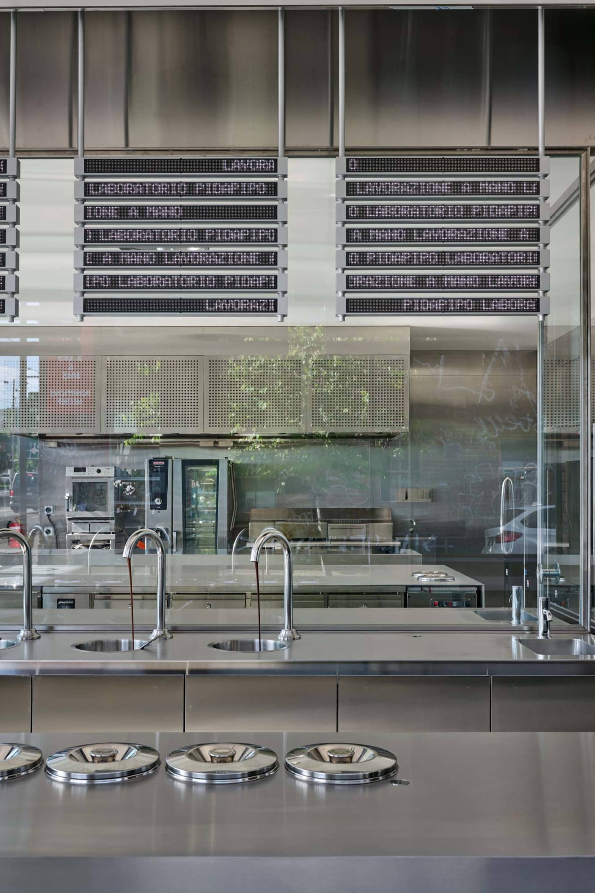 A modern industrial restaurant interior featuring a metal and glass bar counter with digital signs.