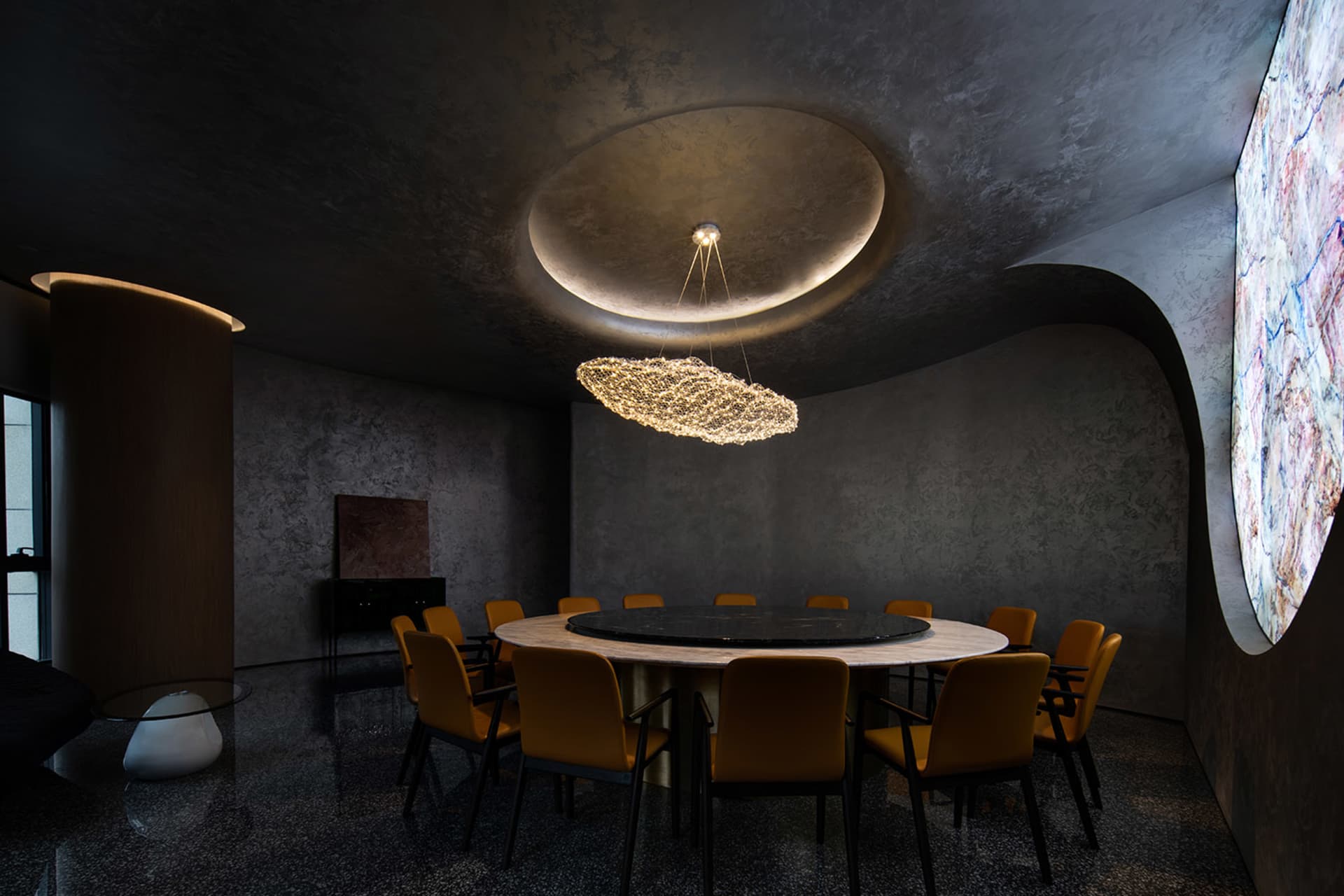 Modern conference room featuring a round table surrounded by chairs, with a chandelier, marble floor, and concrete walls.