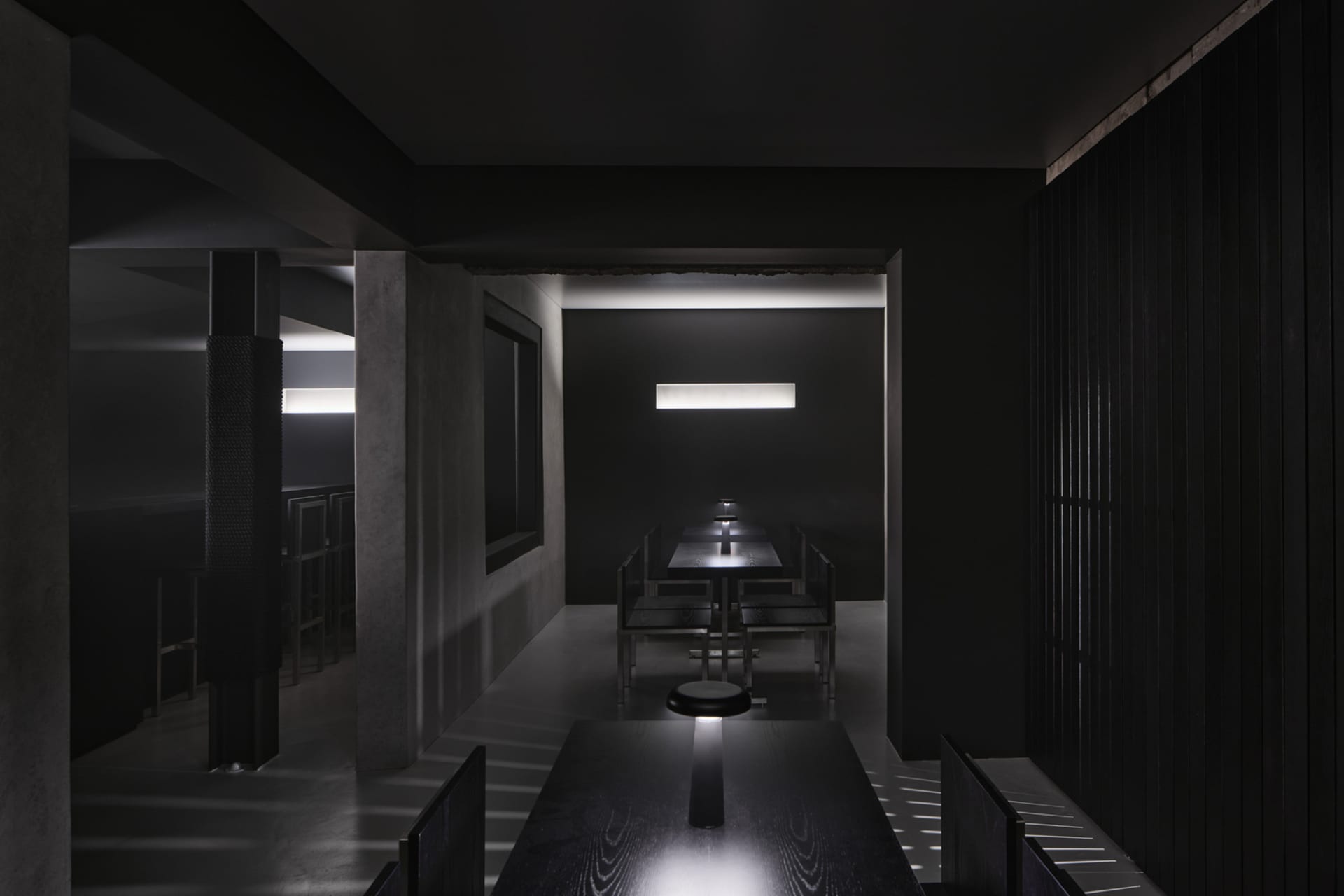 A minimalist restaurant interior with black walls and tables, illuminated by a single light source.