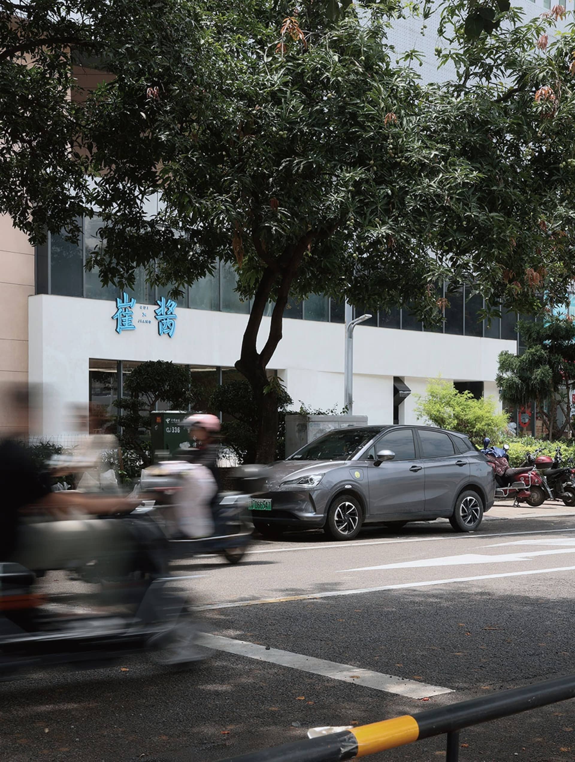 現代都市景觀，灰色汽車停在樹下，背景有其他車輛與建築物。
