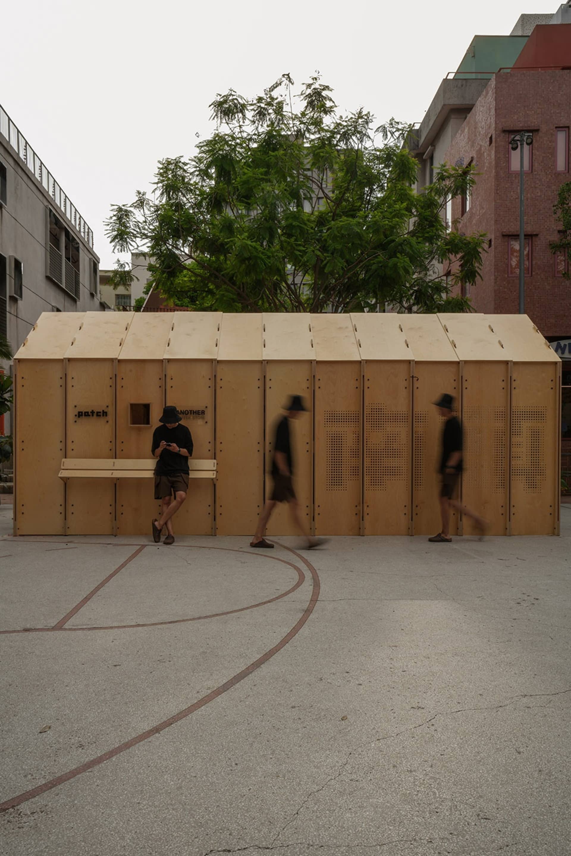 A minimalist wooden shelter in an urban setting, with people walking past.