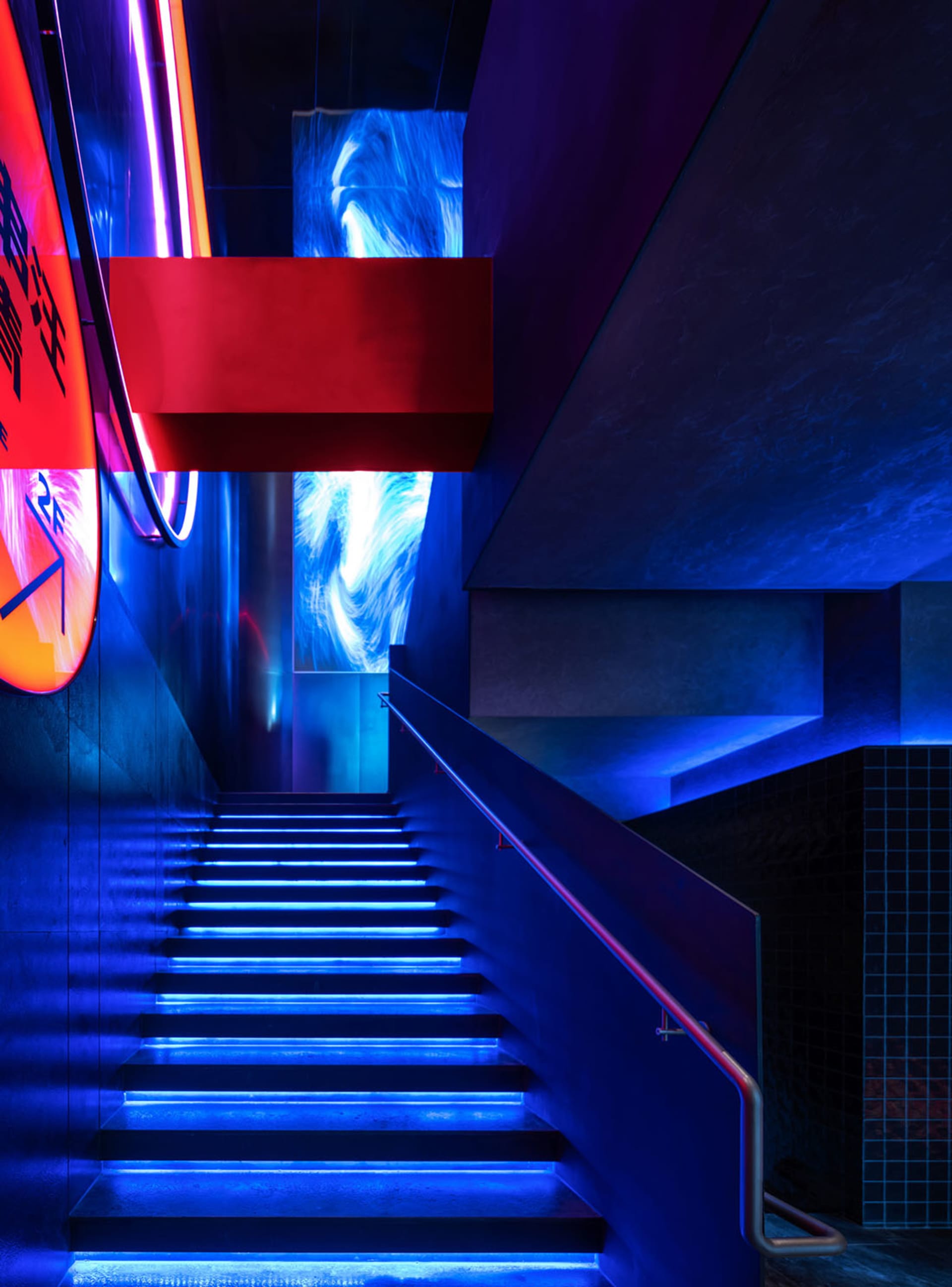 A modern staircase with concrete steps illuminated by blue lights, featuring industrial lighting and a red structural element