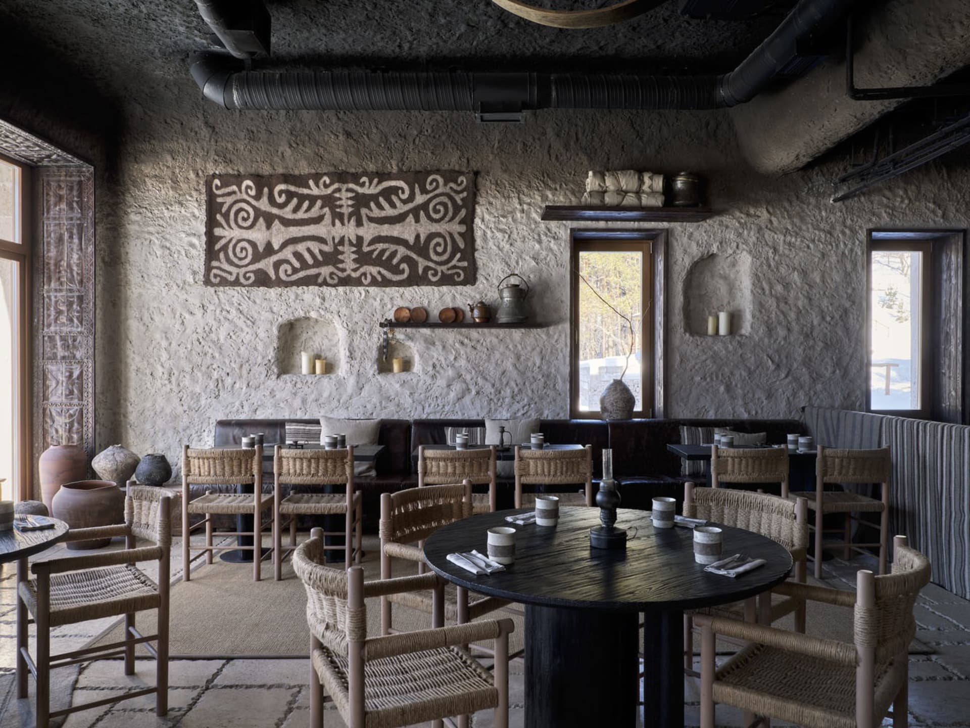 A cozy restaurant interior made of clay, wood, and concrete materials with woven chairs and round tables.