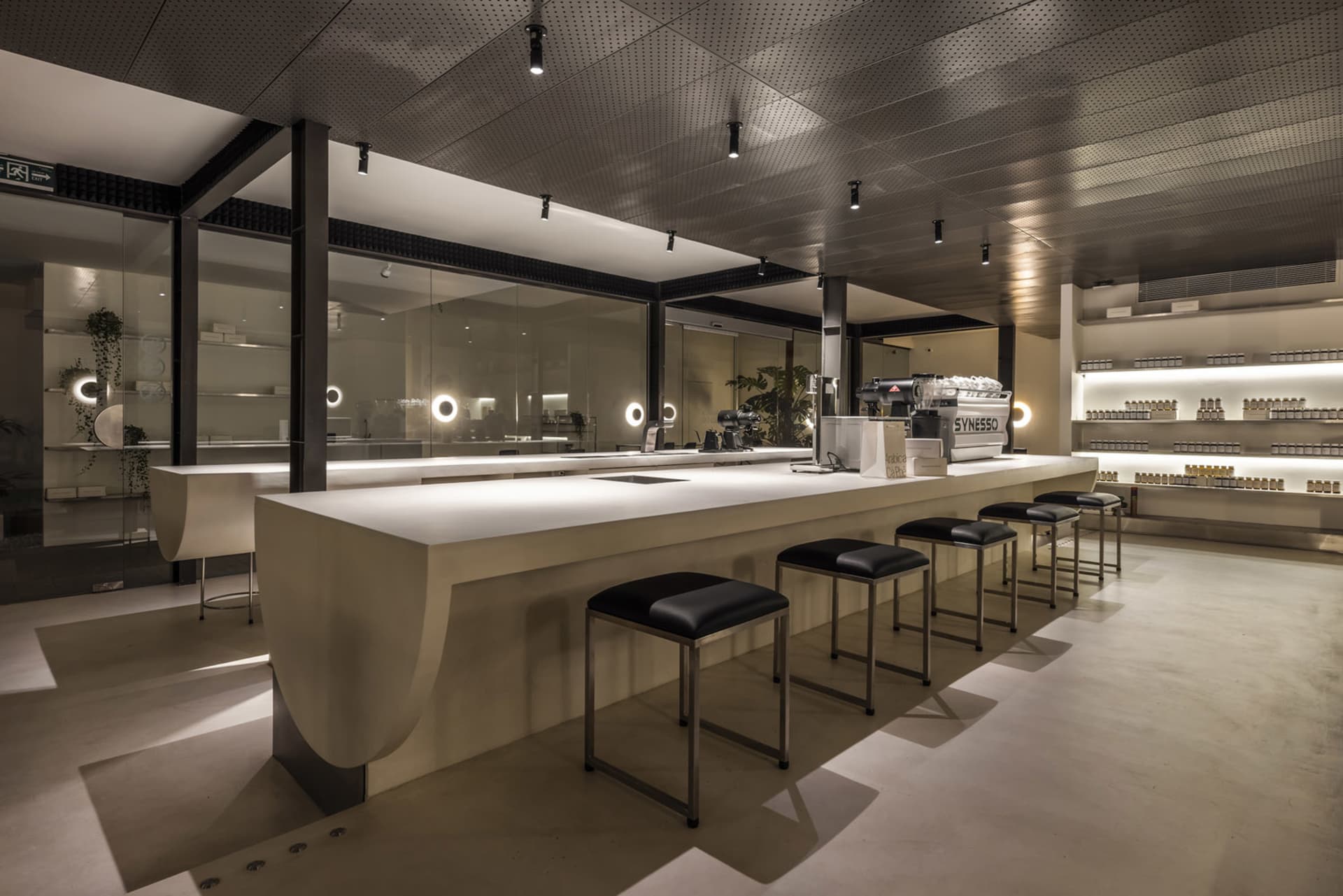 A modern coffee shop with a minimalist aesthetic, featuring a concrete bar counter, black stools, and white walls with floor-