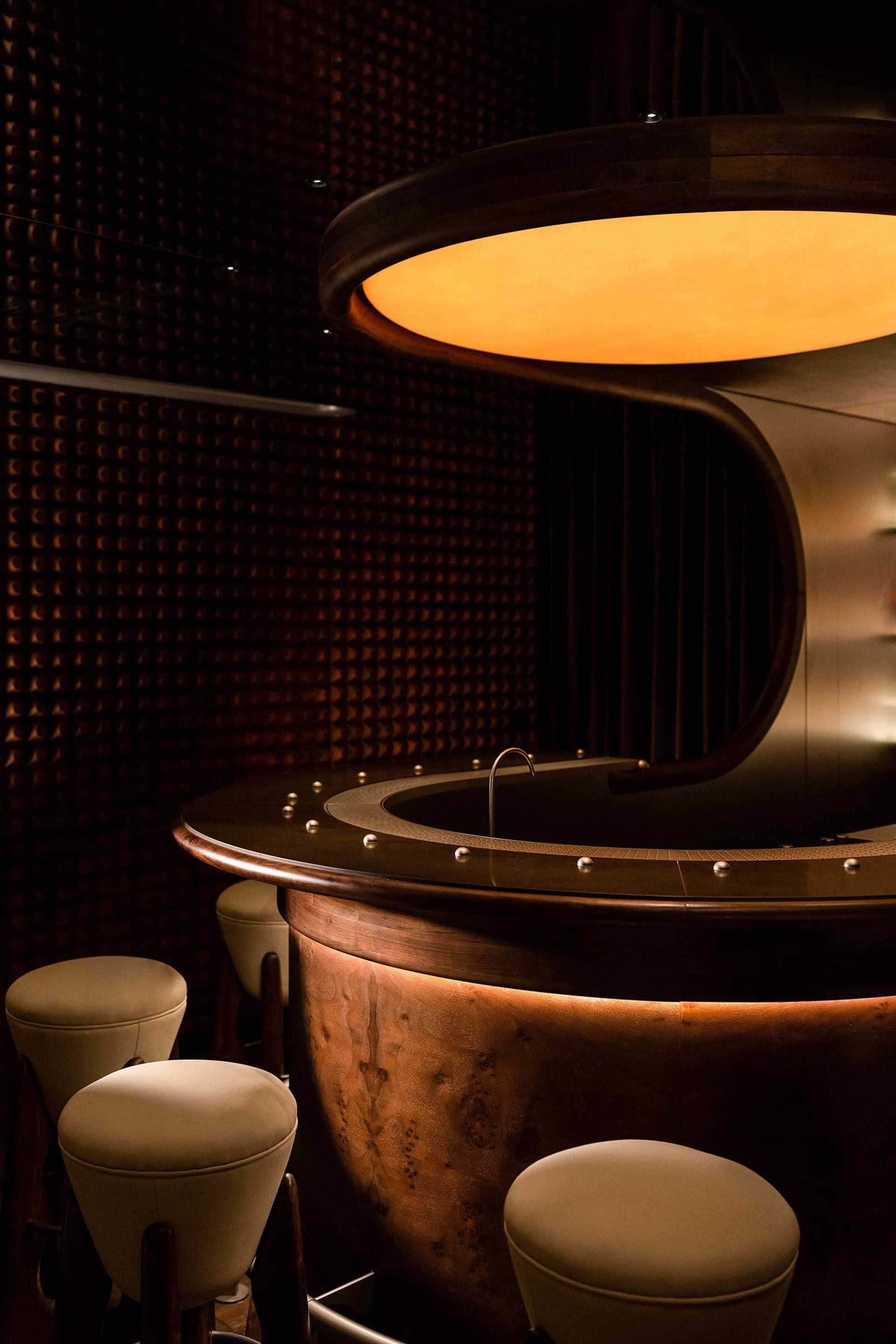 A modern industrial bar with metal barstools against a dark, textured wall, illuminated by warm lighting above.
