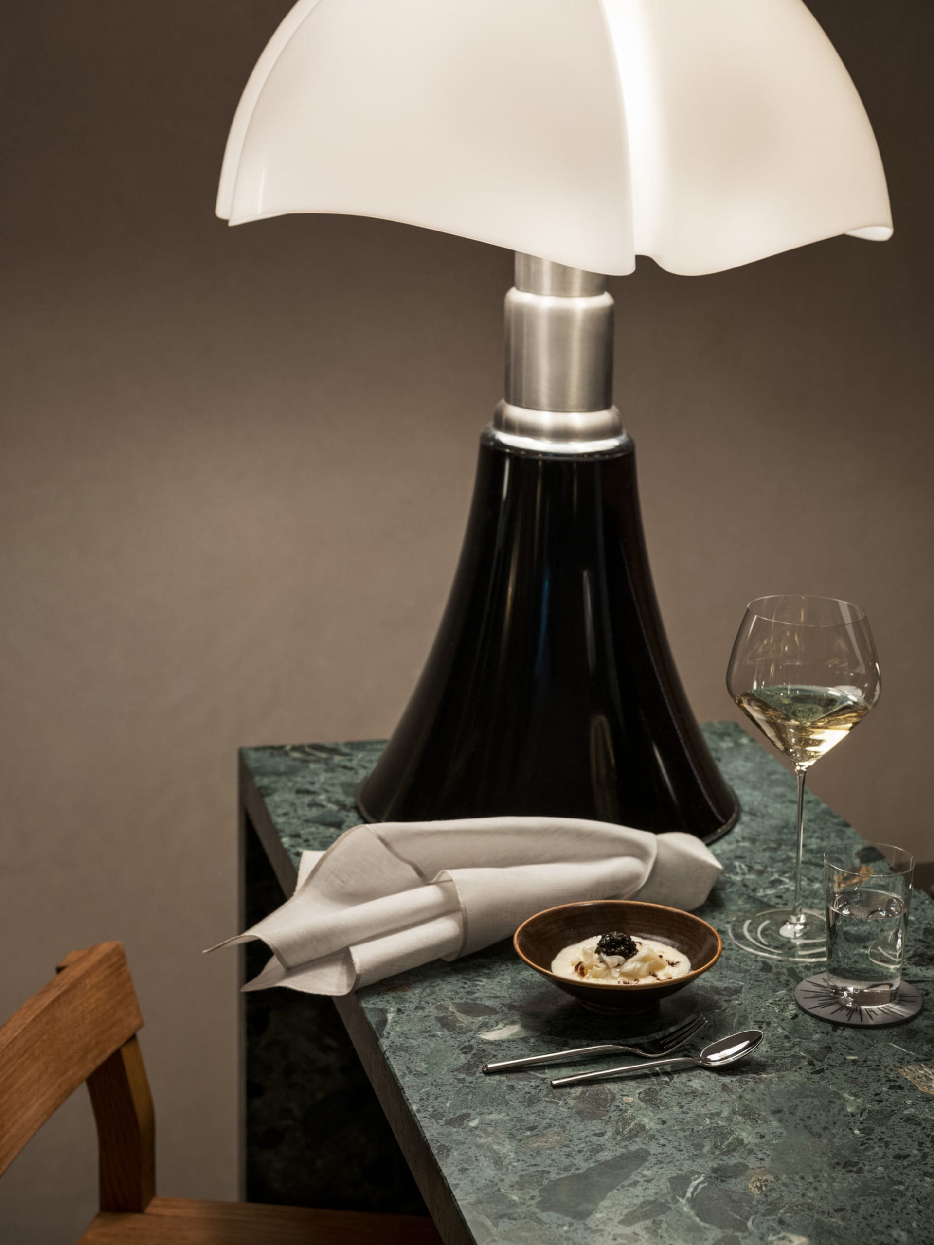 A modern restaurant table setting featuring a green marble table, a white shaded table lamp, a glass of white wine, a bowl of