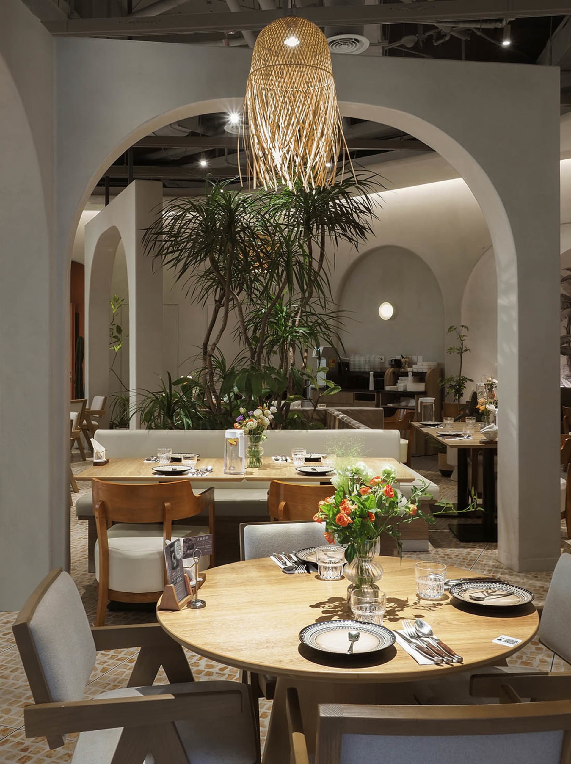 Modern restaurant interior with wooden tables, glass vases, and plush fabric chairs.