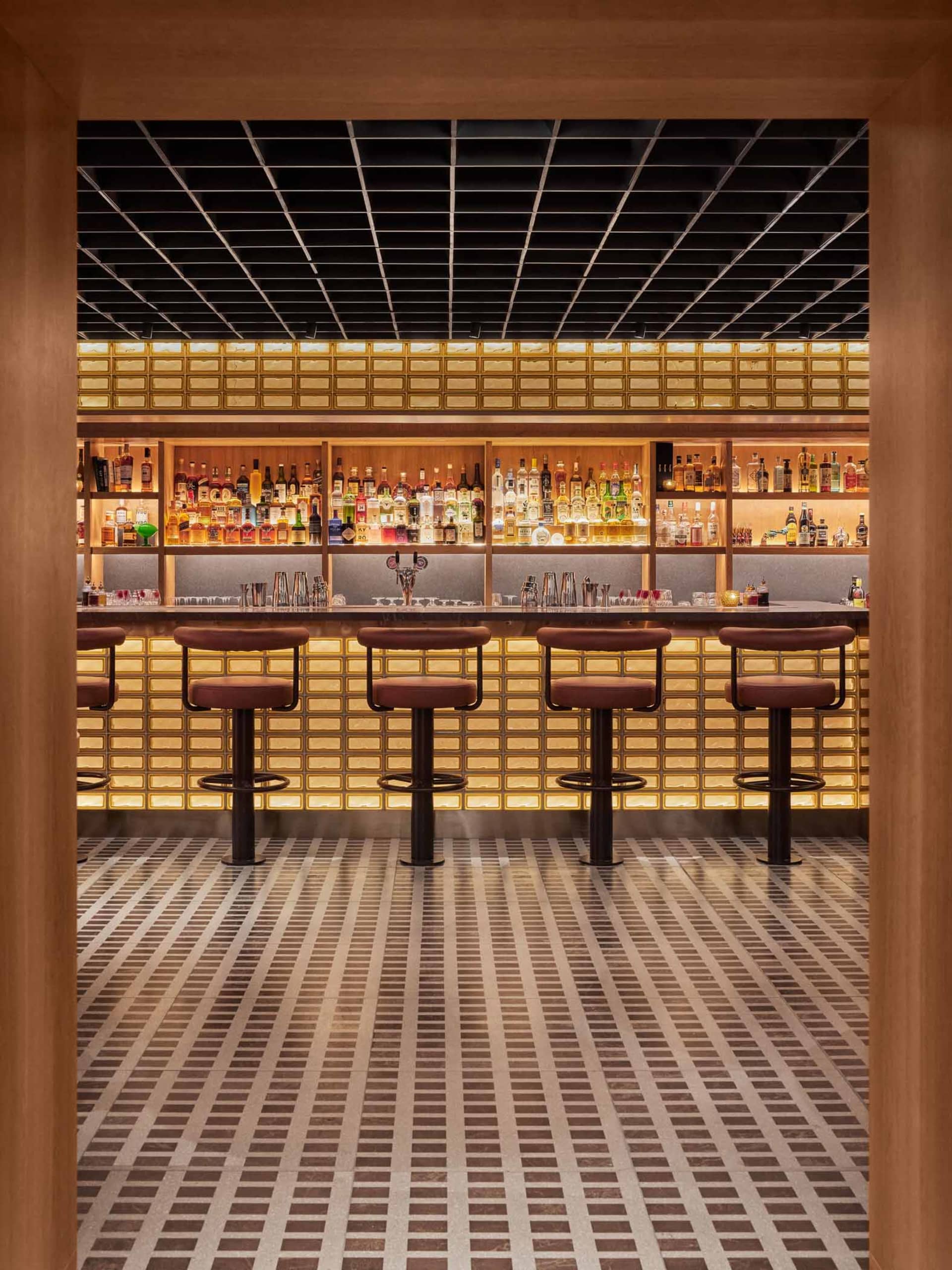 Modern bar with tiled floor, stools, and backlit shelves filled with various bottles of alcohol.