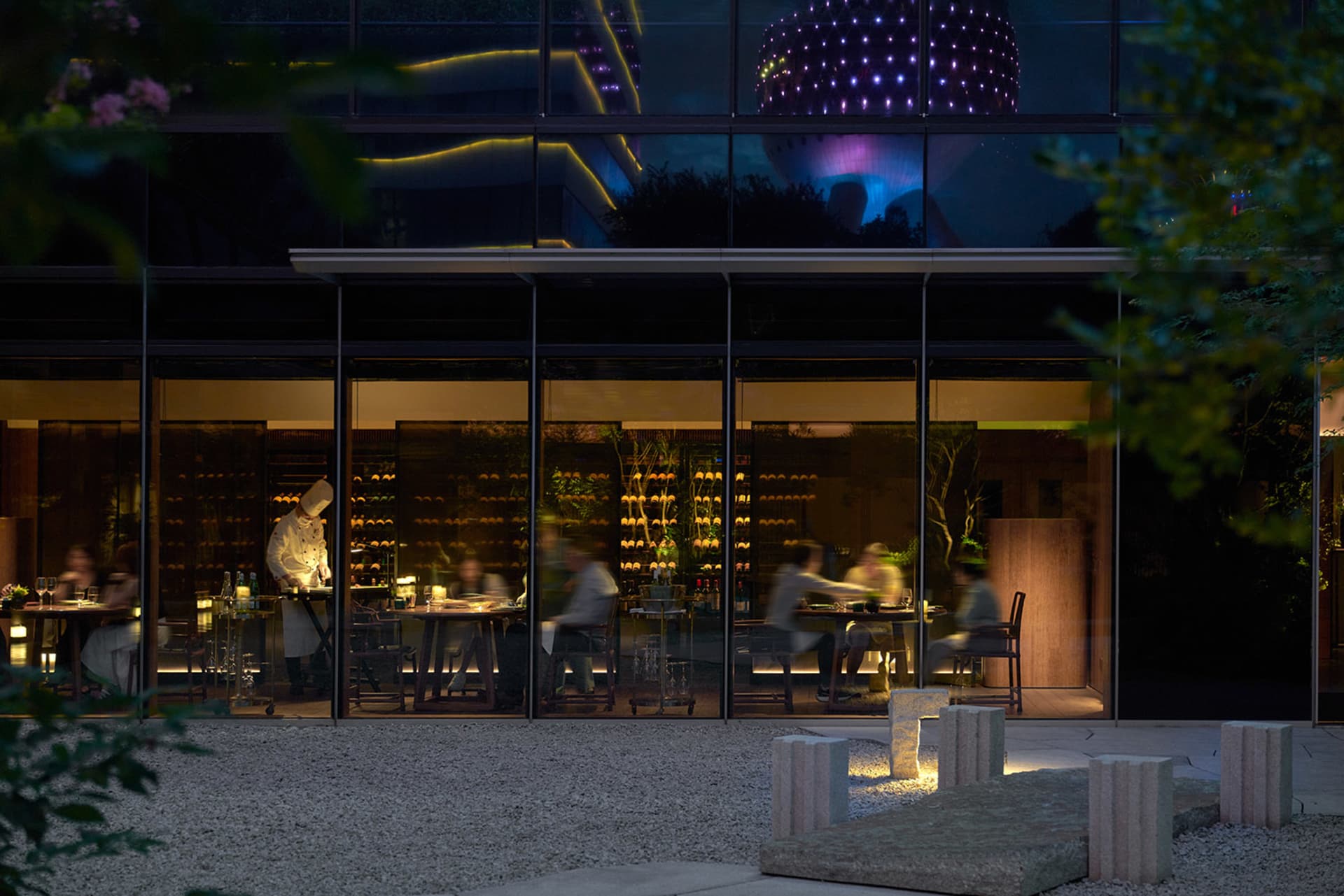 A modern restaurant exterior at night with a glass facade reflecting city lights and a chef working inside.