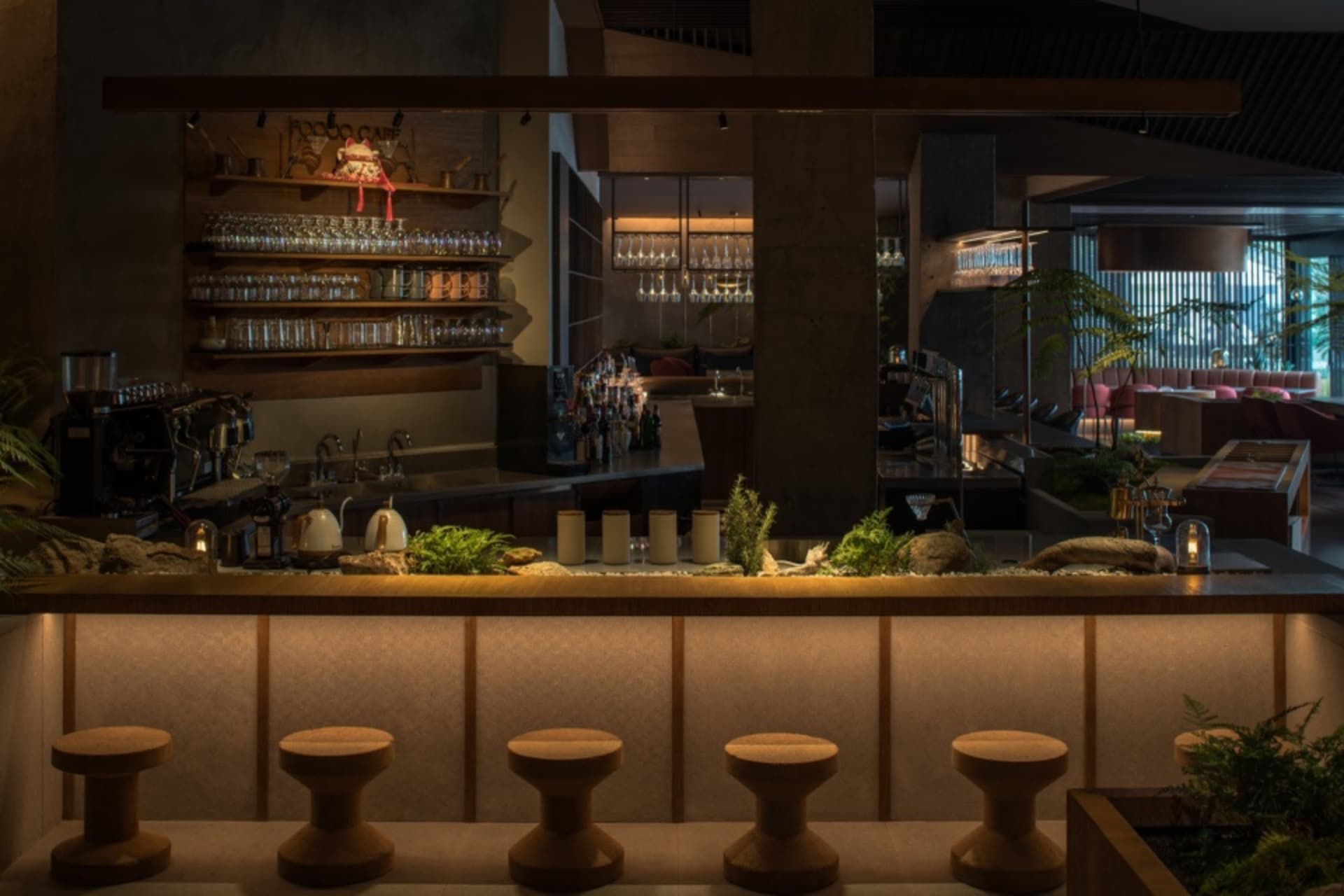 A modern bar interior featuring wooden stools and concrete columns, with a well-stocked bar counter and greenery on the count