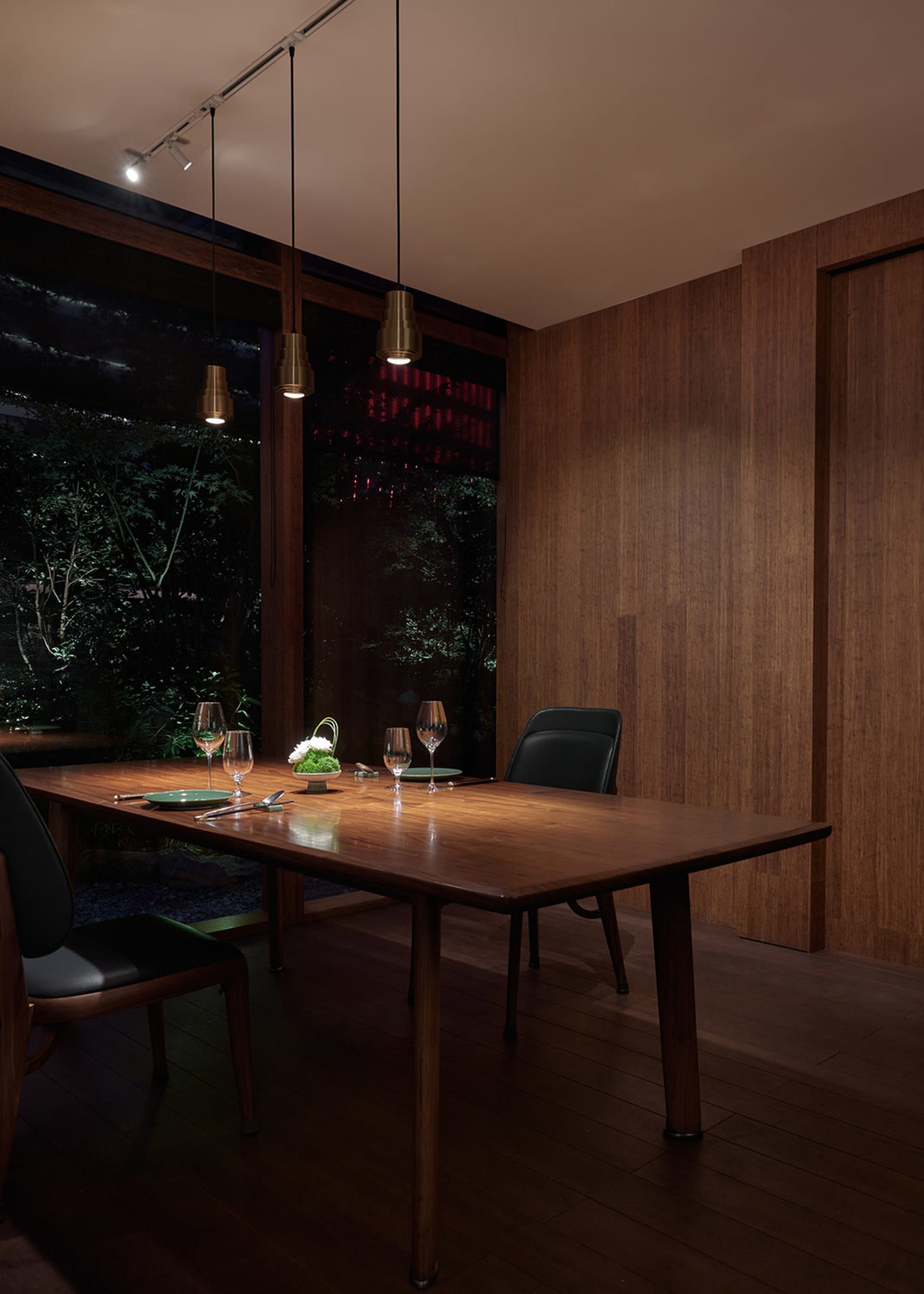 Modern dining room with wooden table, chairs, and pendant lights, featuring a view of greenery outside through large windows.