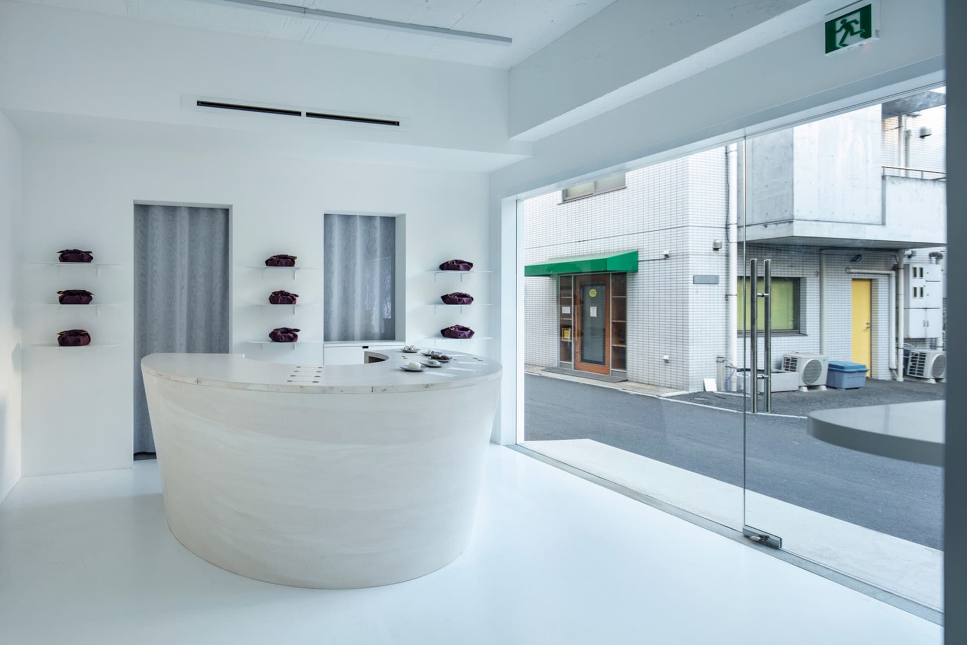 A minimalist retail storefront with a large, curved white marble reception desk and purple bags displayed on shelves against