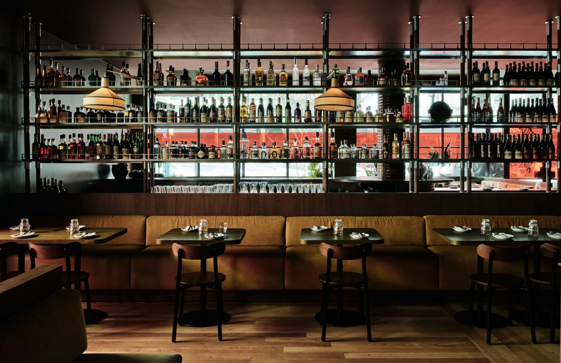 Modern restaurant bar interior with wooden tables, metal stools, and shelves filled with bottles.