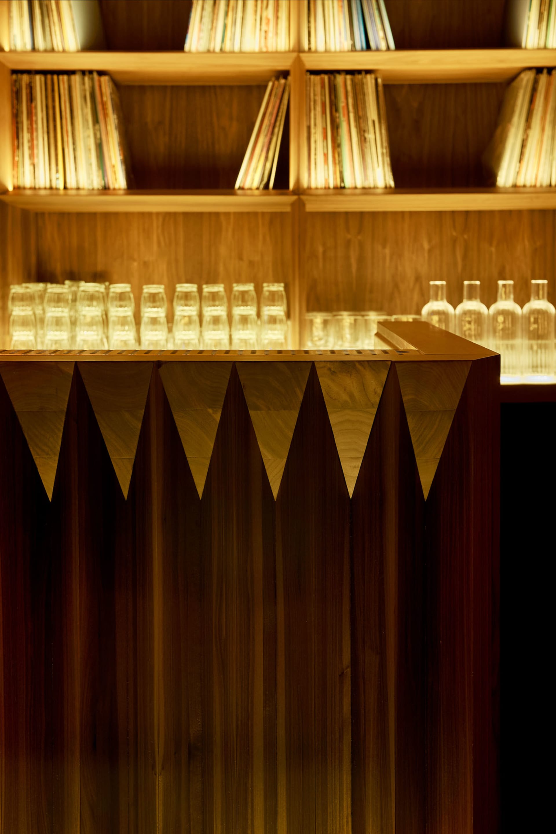 A modern bar counter made of wood, with shelves behind it containing records and glasses.
