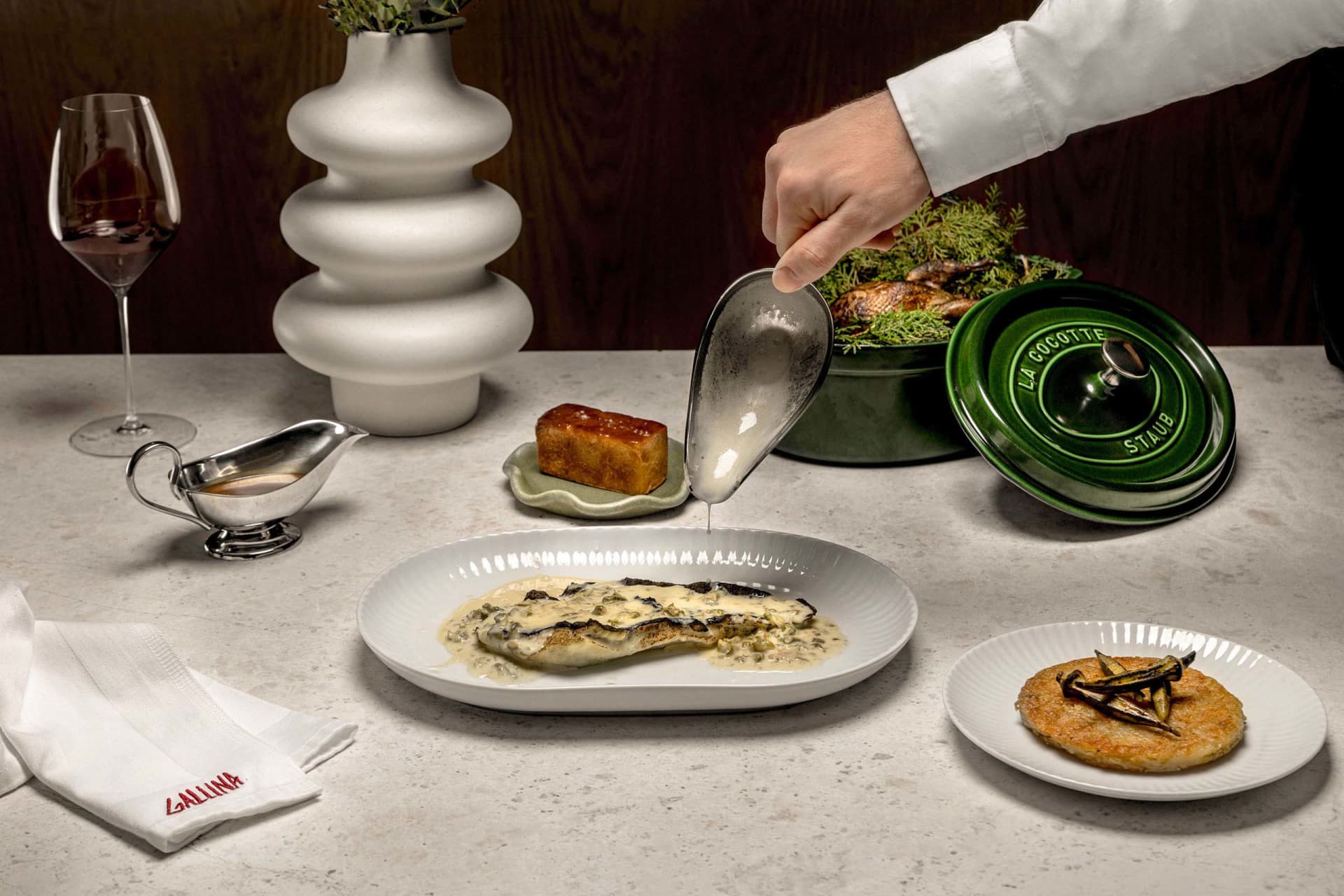 A modern food service setting featuring ceramic dishware, a green cocotte dish, and a glass of wine.