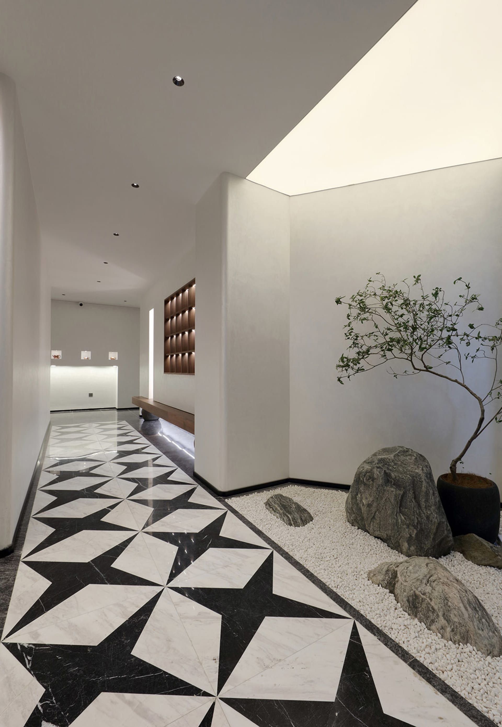 A modern entryway featuring black and white marble flooring, a small rock garden with a tree, and a long wooden bench against