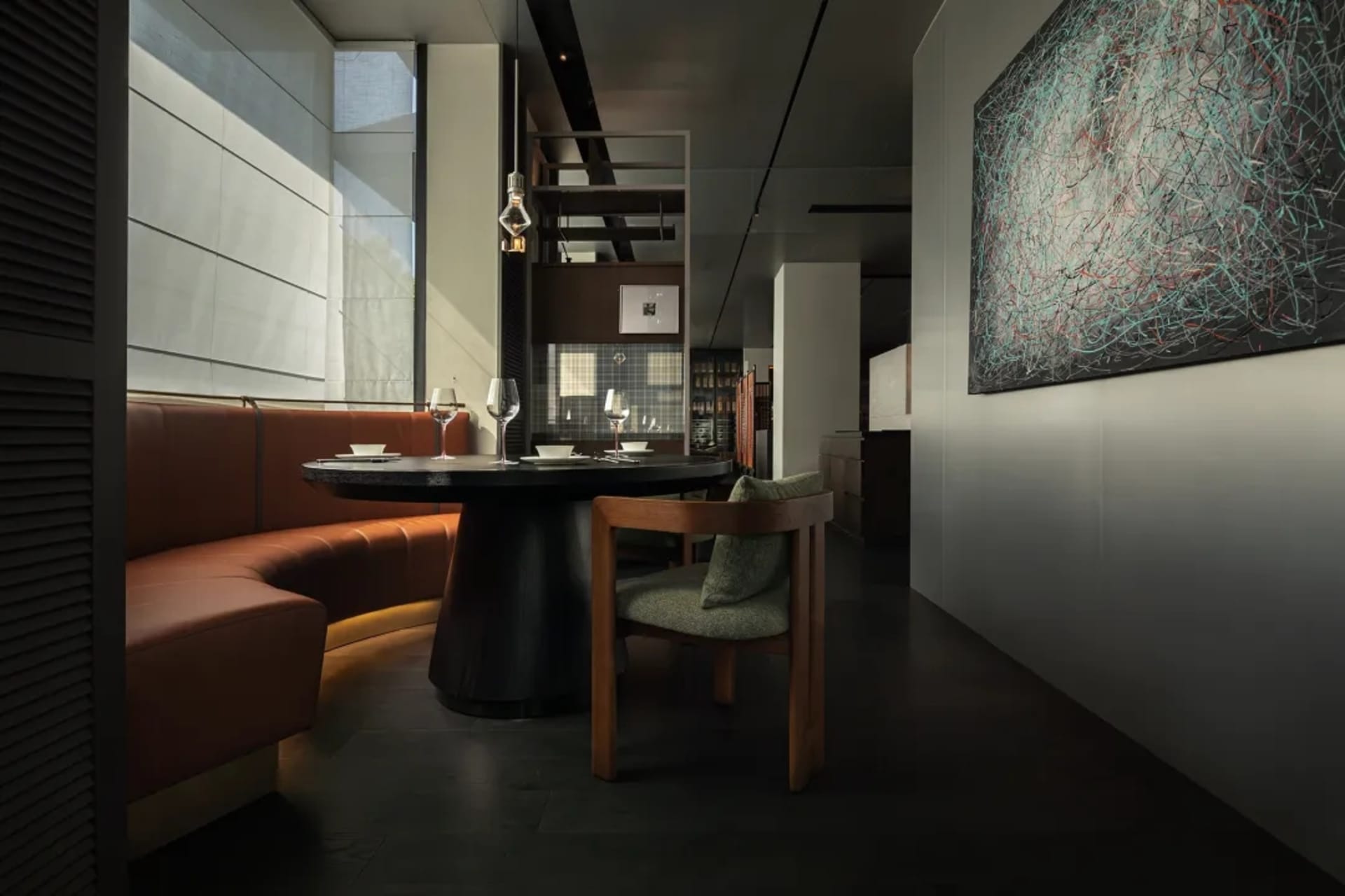 A modern restaurant interior featuring a round dining table with leather seating, a large abstract painting, and dark wooden