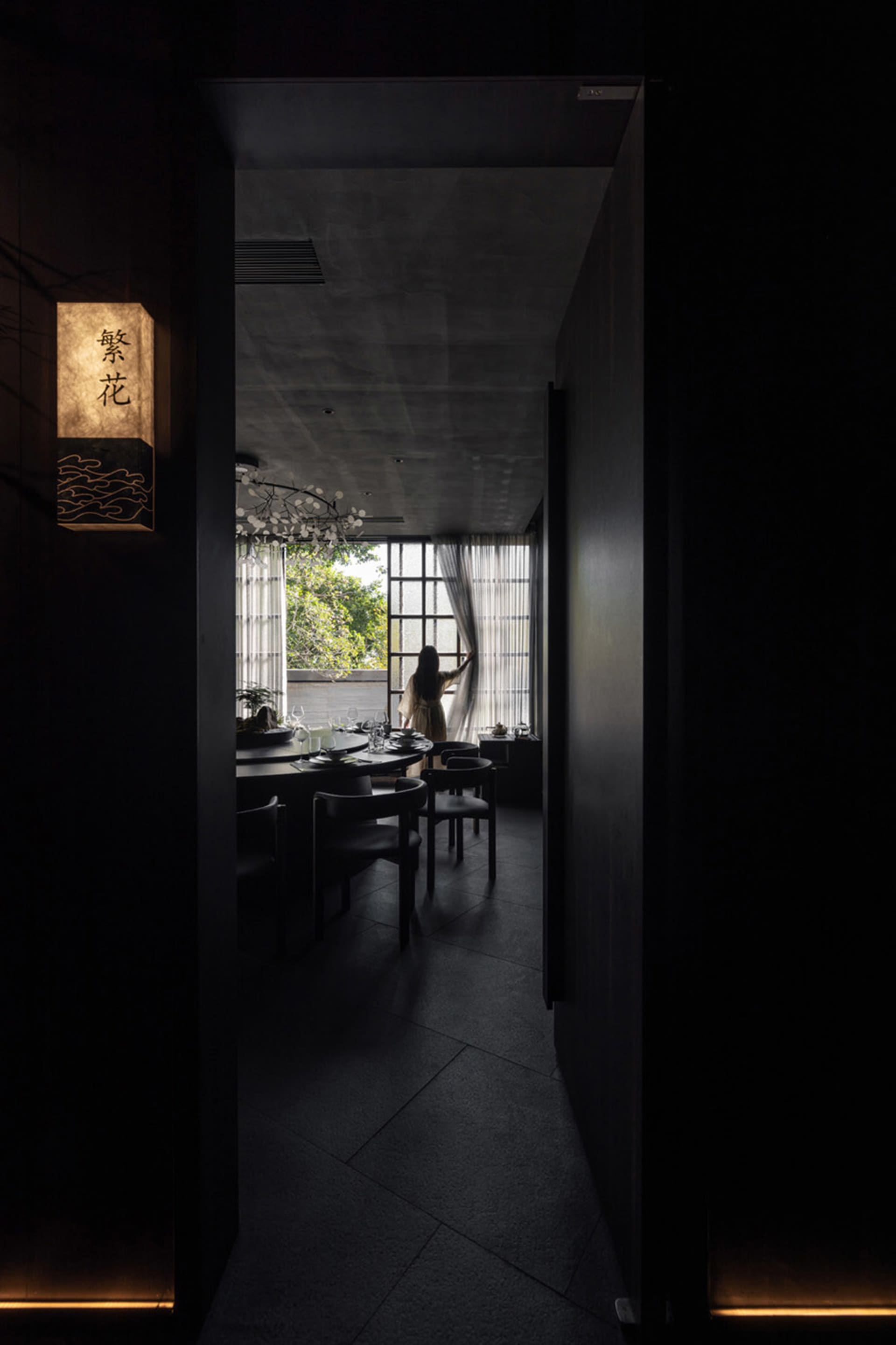 極簡風格的餐廳，混凝土牆面與地板，配有圓桌和椅子，一人正在調整窗簾。