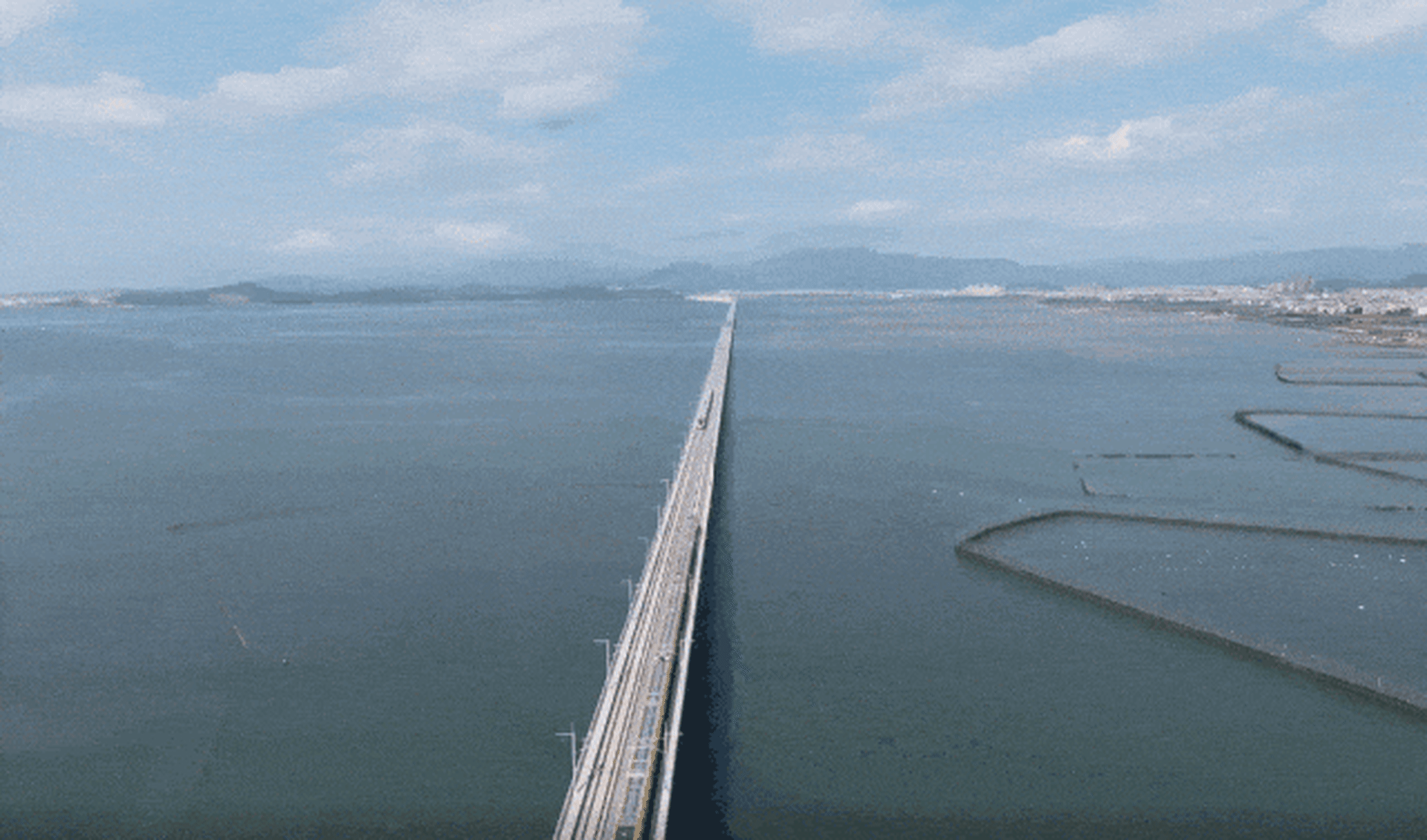 A steel bridge extending across a wide expanse of water with a cloudy sky and distant hills in the background.