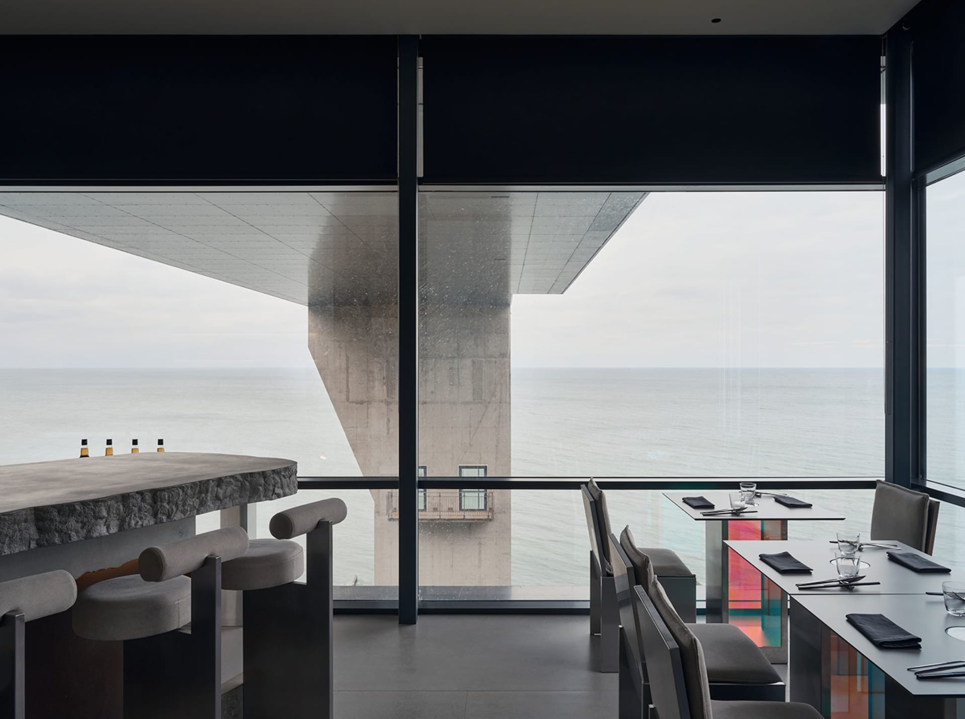 Interior of a modern restaurant bar featuring a concrete countertop, glass windows overlooking the ocean, and metal chairs wi
