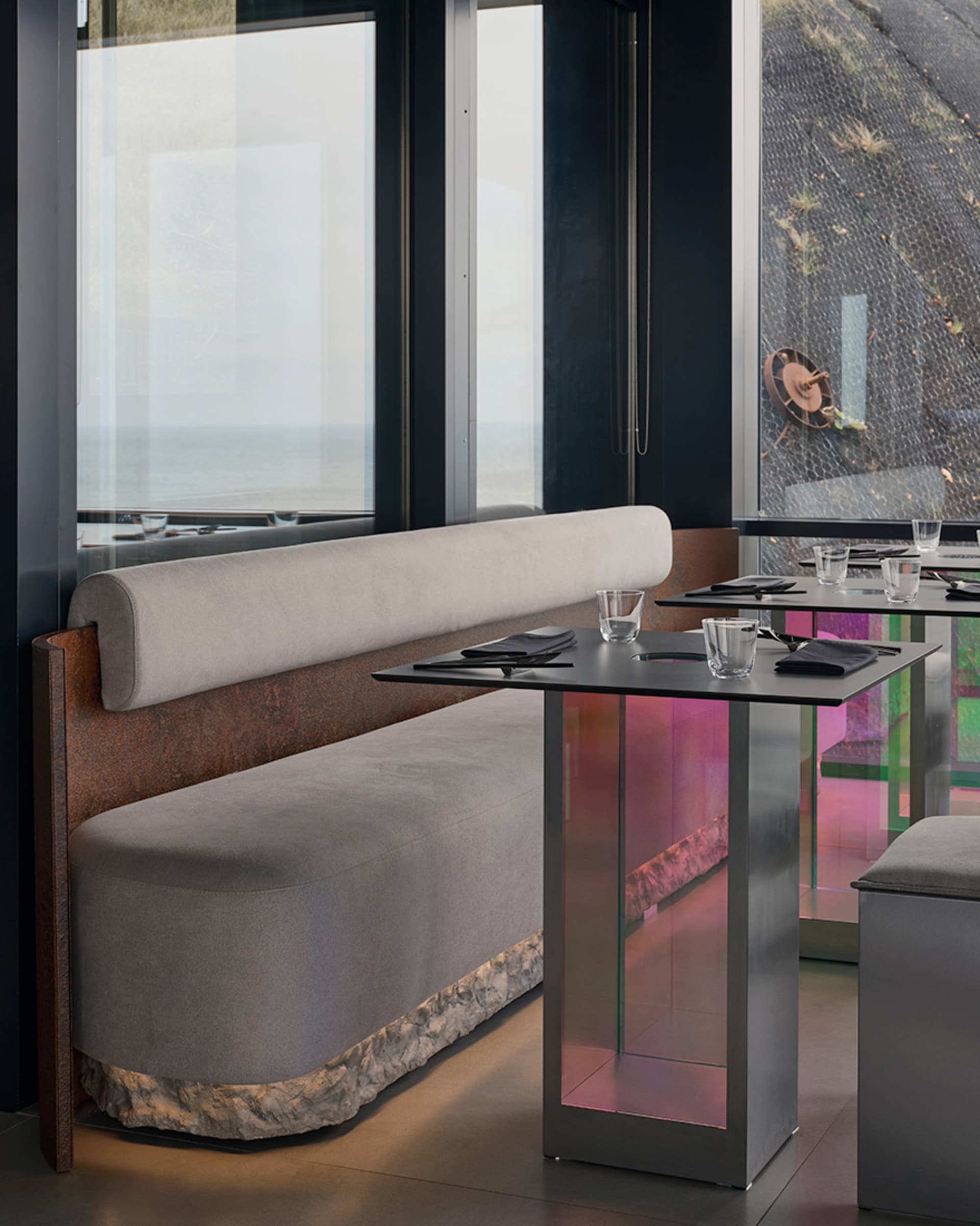 Modern restaurant interior with metal tables, fabric booth seating, and glass elements.