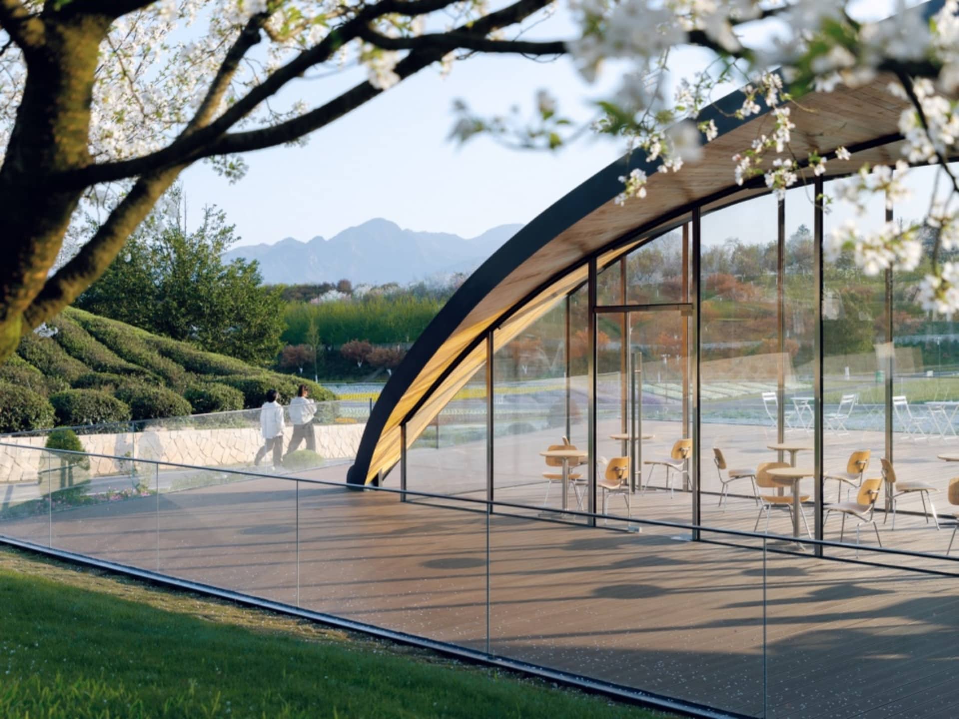 Modern glass terrace with wooden seating and metal railings overlooking greenery and hills.