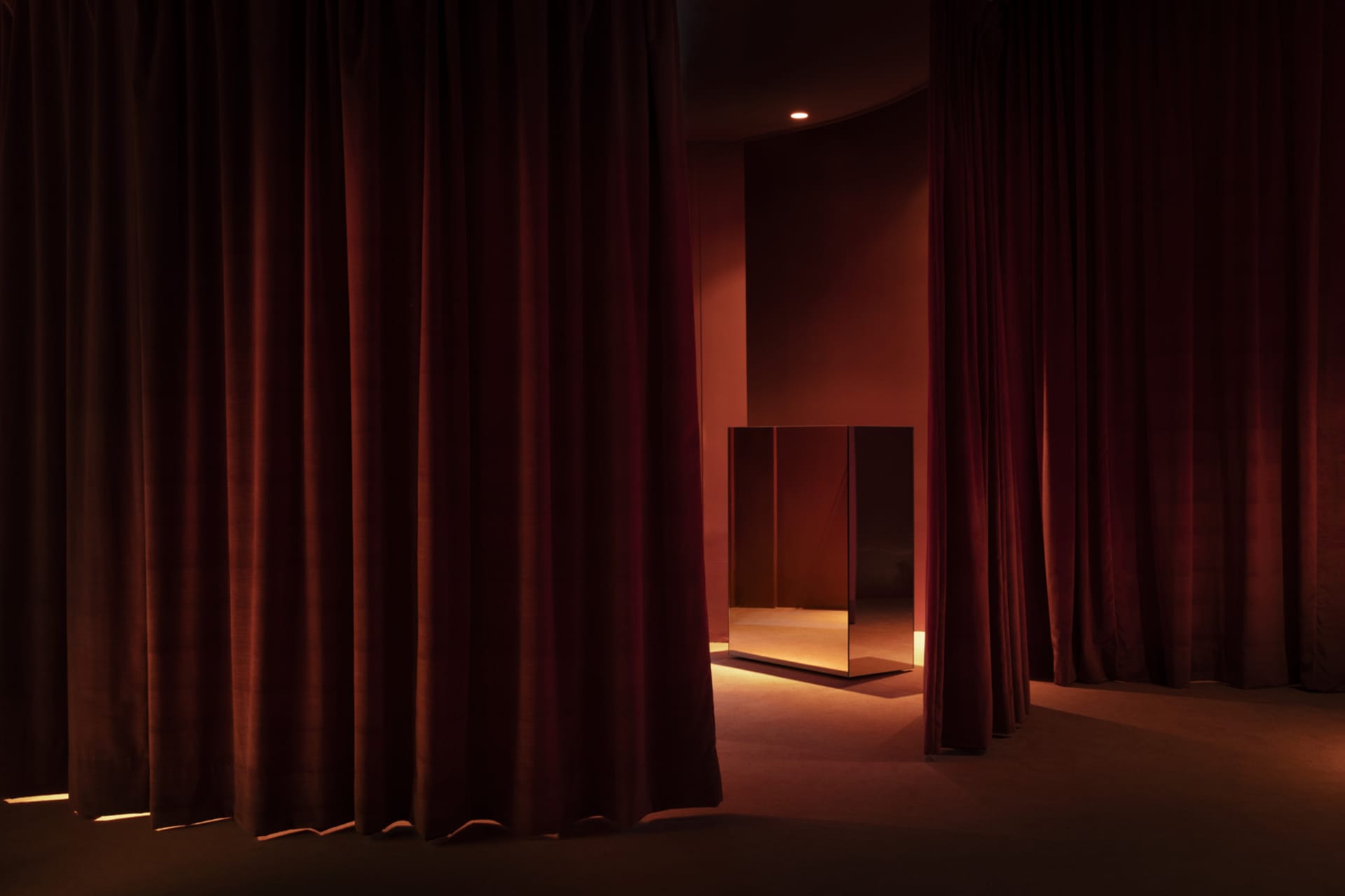 Dark red velvet theater stage curtains in front of a dimly lit, minimalist interior with a mirrored box in the center.