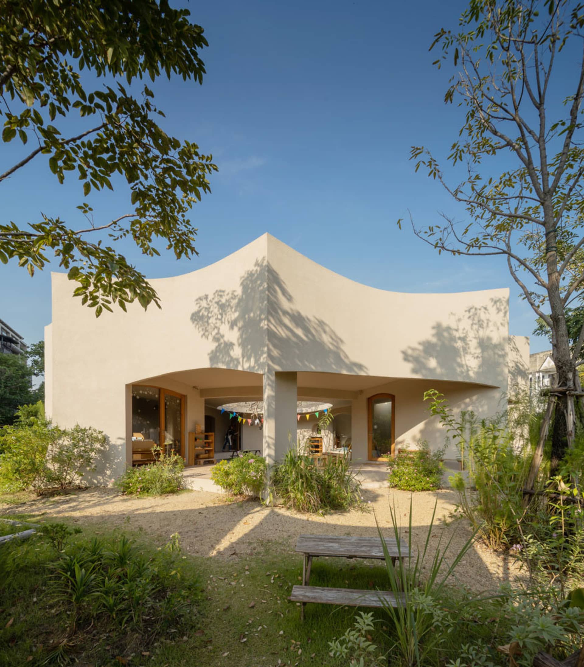 A modern house with a curved roof and concrete walls surrounded by greenery and a wooden bench in the foreground.