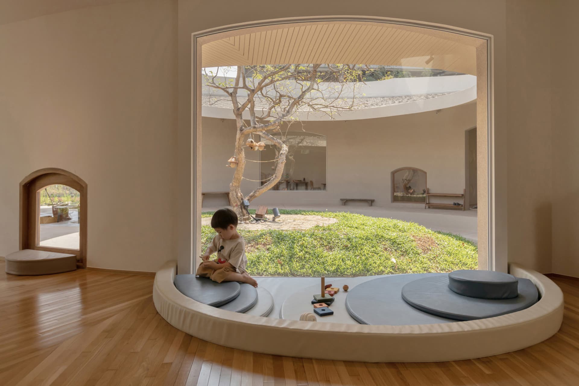A child plays on a circular platform with soft furnishings in a minimalist playroom with a tree in the background.