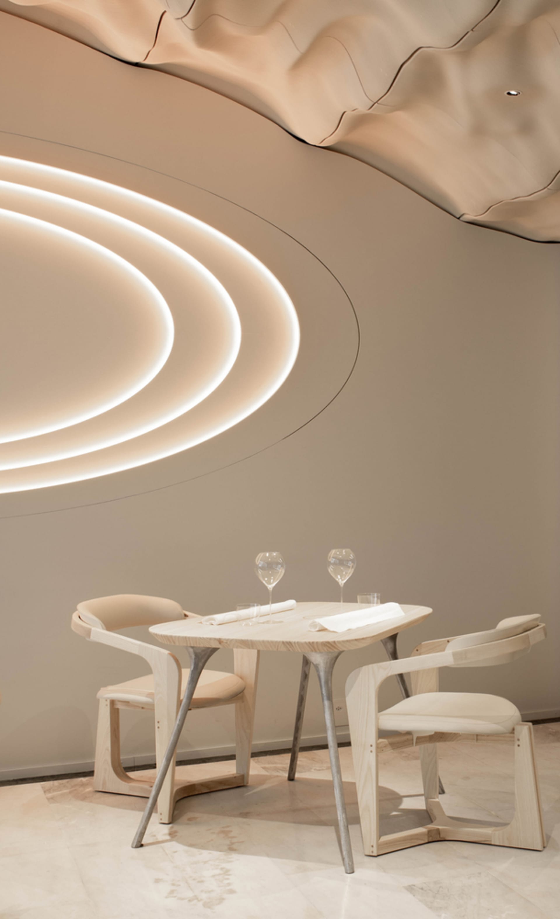 Modern restaurant interior with round wooden table, chairs, and illuminated architectural design on the ceiling.