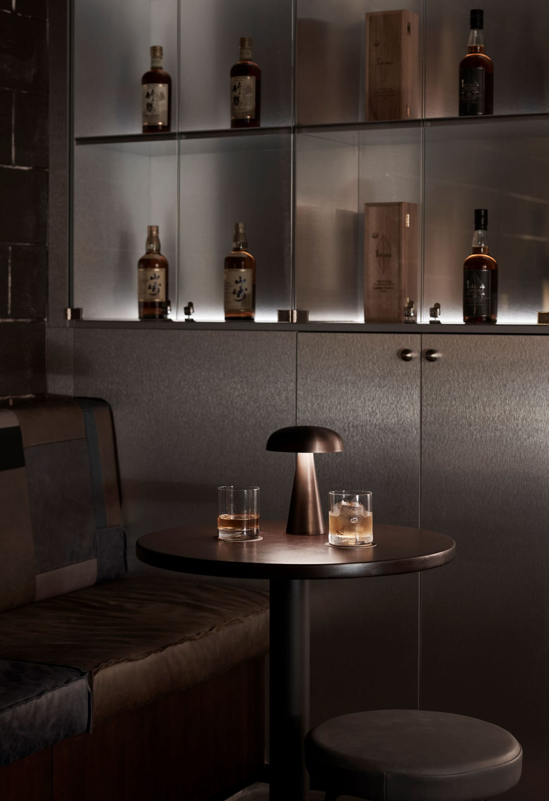 A bar counter with a metal finish, featuring a round table with two glasses of whiskey and a small lamp.