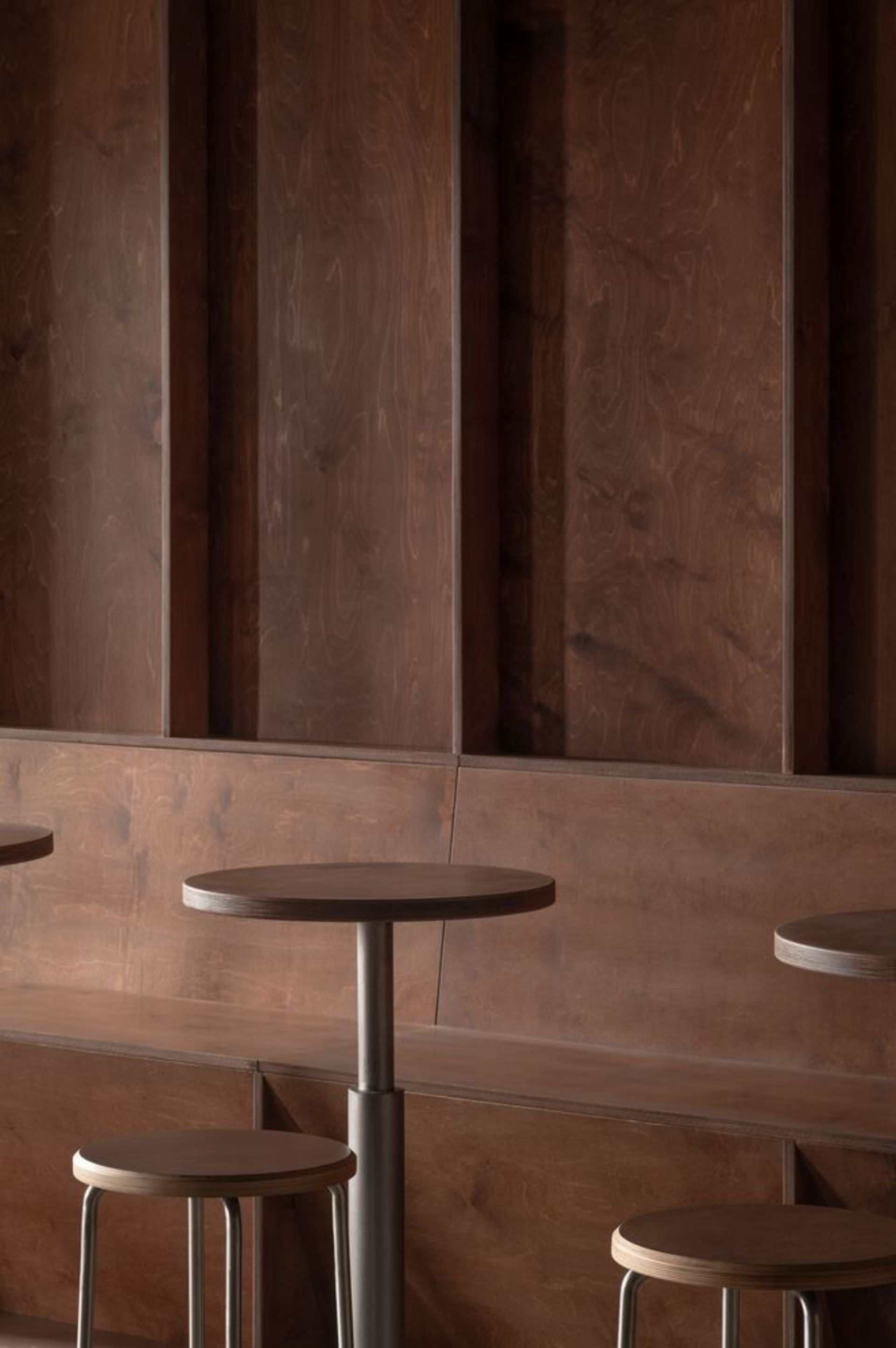 Modern café seating area featuring wooden stools and a round table with a concrete base, set against a wooden paneled wall.