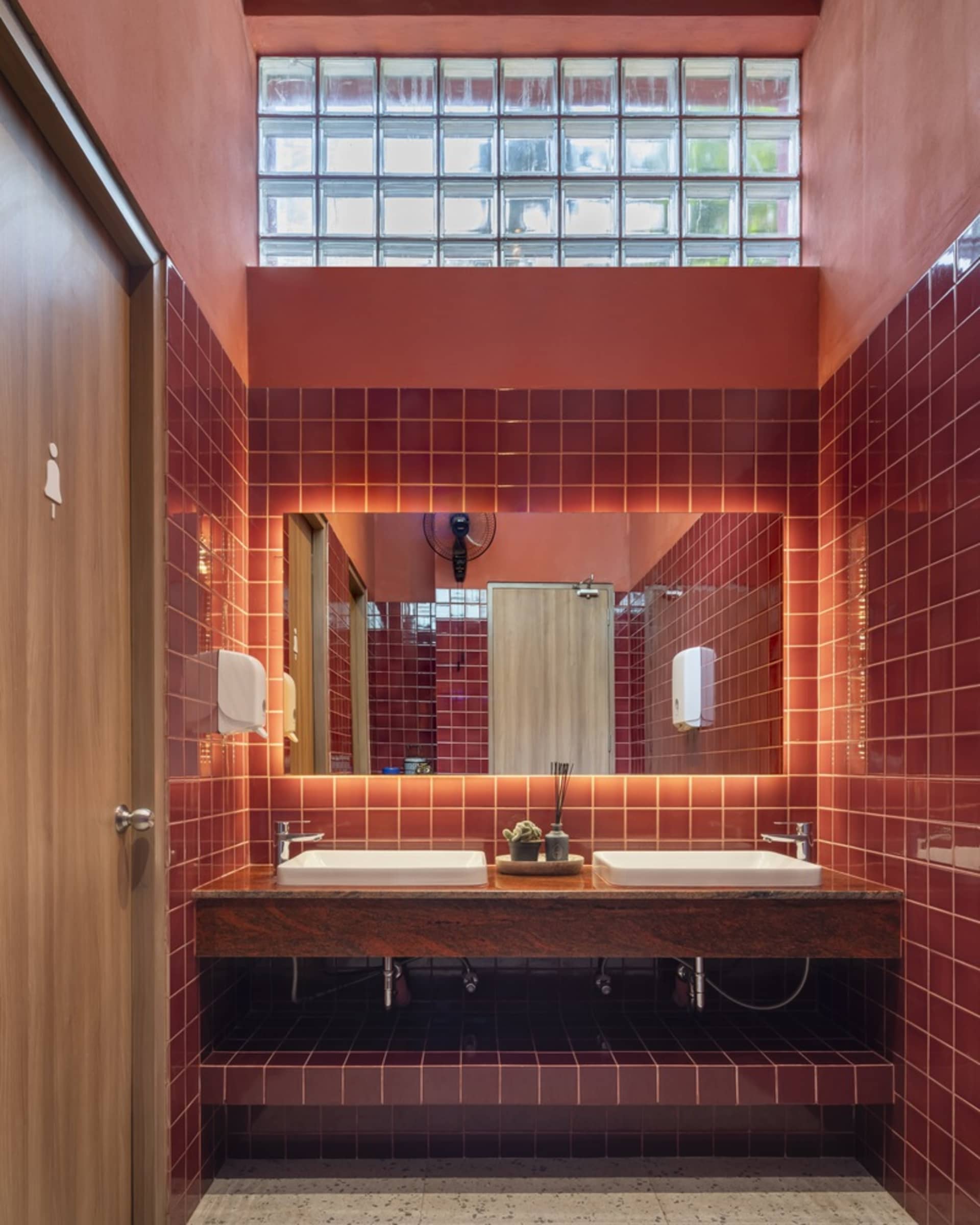A modern public bathroom with red tiled walls, glass brick ceiling, and a wooden door.