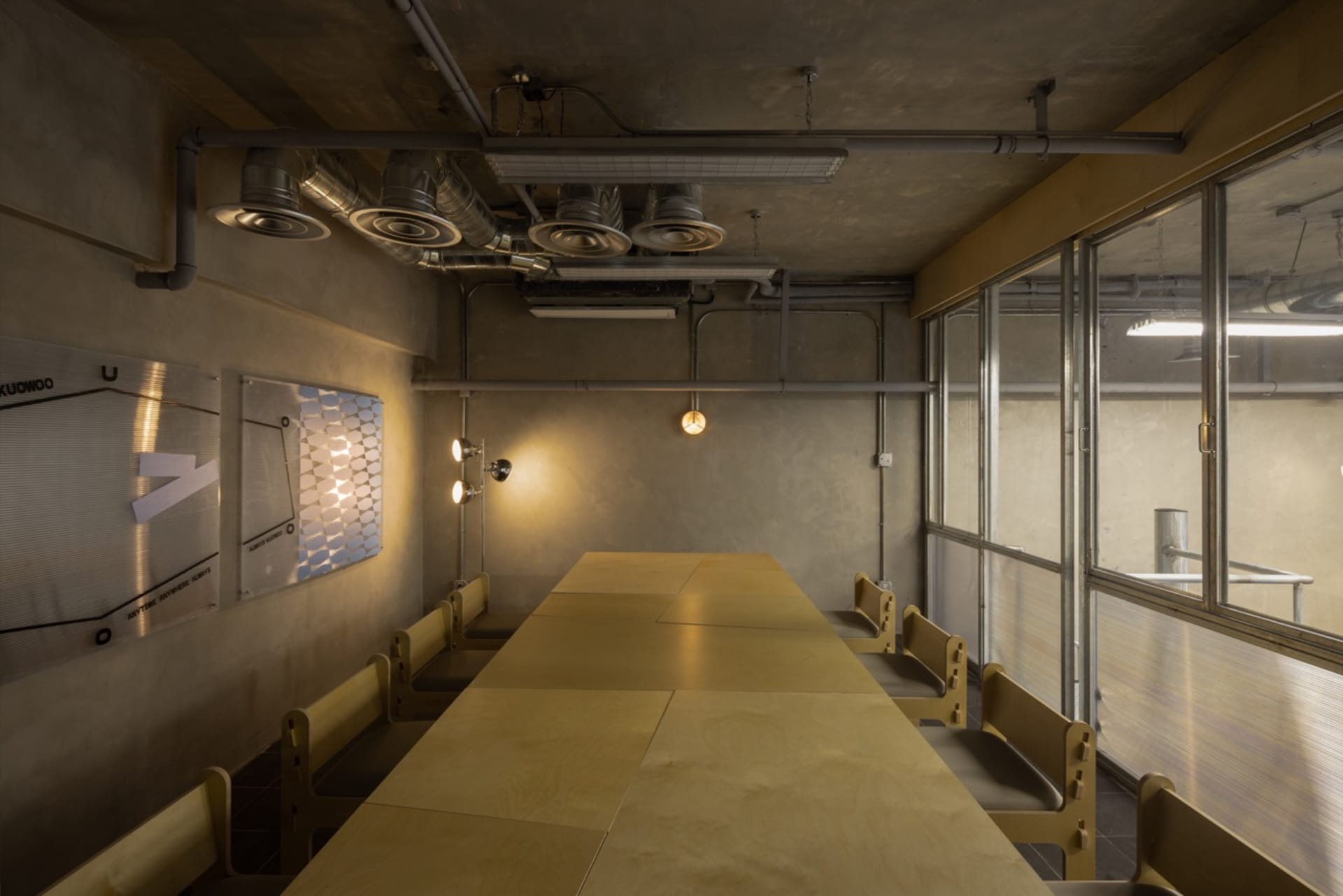 An industrial-style conference room with a long wooden table and wooden chairs, featuring exposed pipes and lighting fixtures