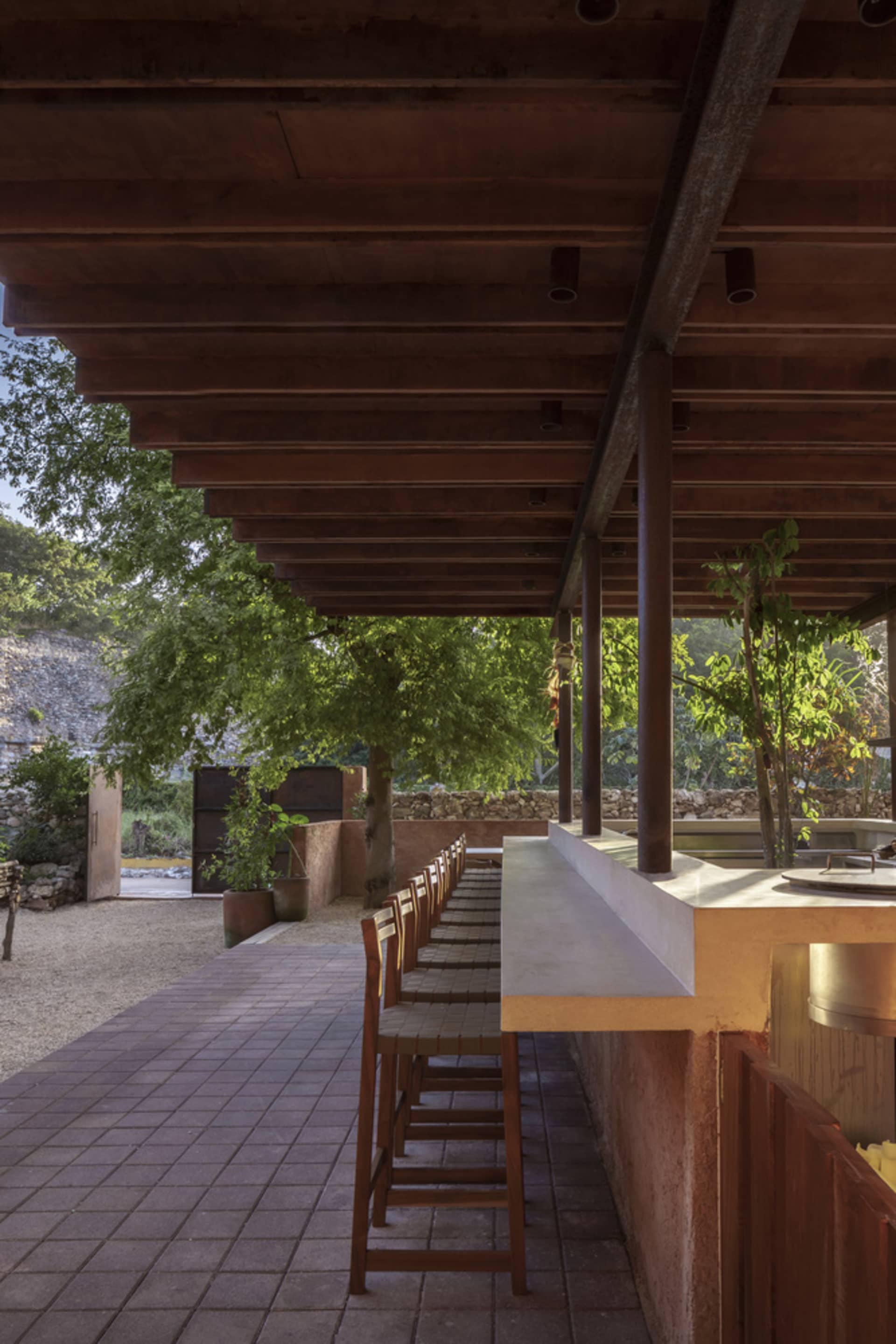 Outdoor patio area with wooden tables, bar stools, and a concrete countertop under a wooden pergola with greenery.