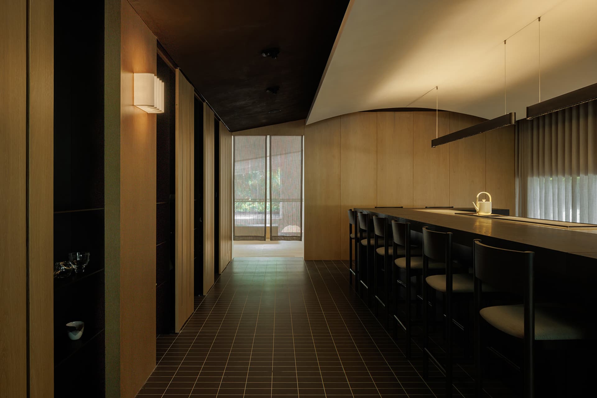 A modern restaurant bar with wooden interior, featuring a long counter and chairs, large windows, and soft lighting.