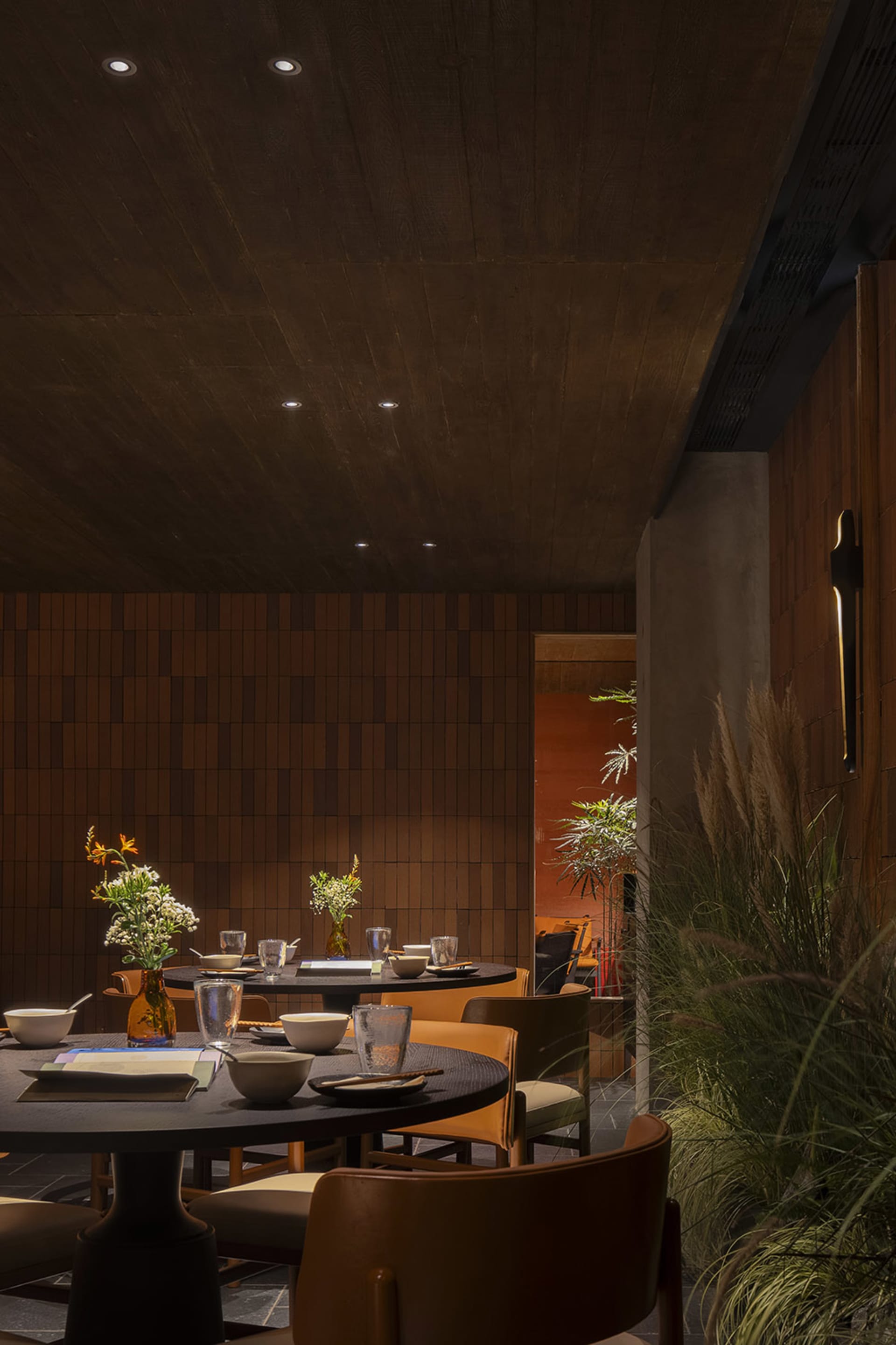 Modern restaurant interior with wooden tables and chairs adorned with plant arrangements, set with bowls, glasses, and books.
