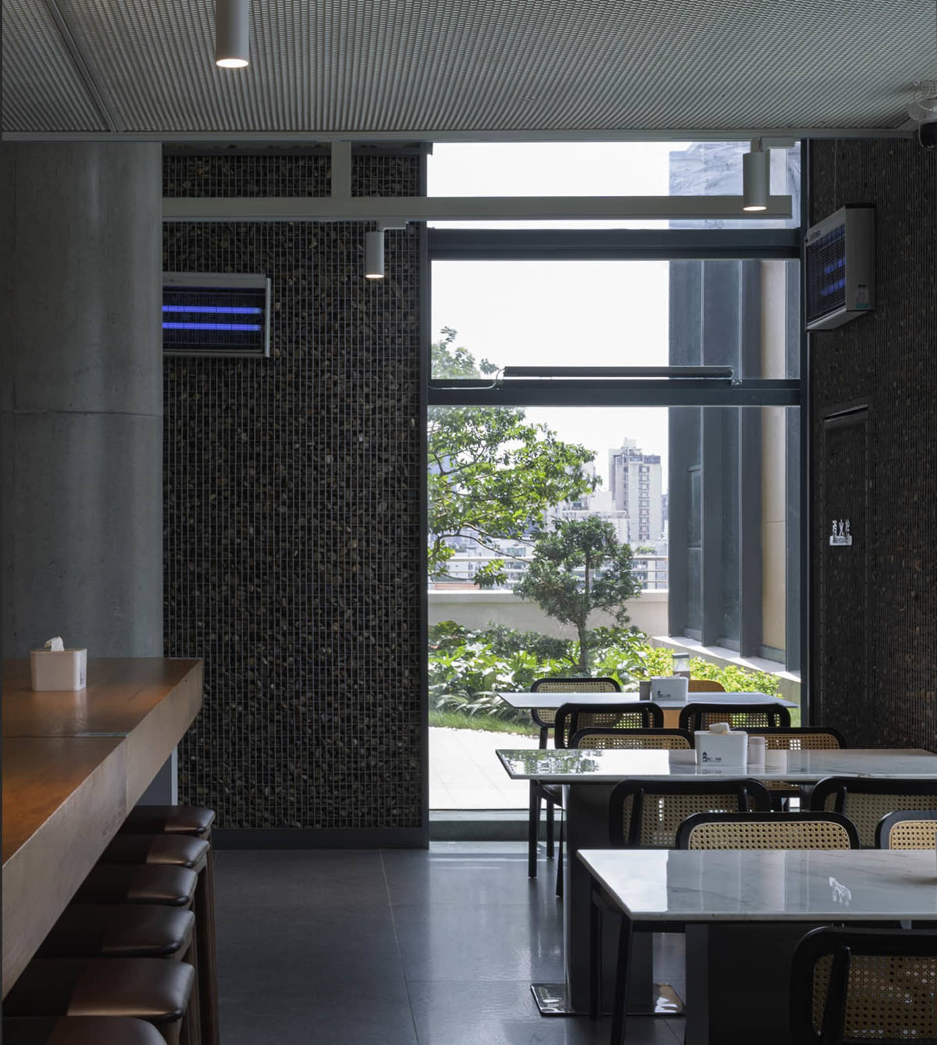 Modern industrial restaurant interior with concrete walls and glass doors, featuring white marble tables and black chairs.