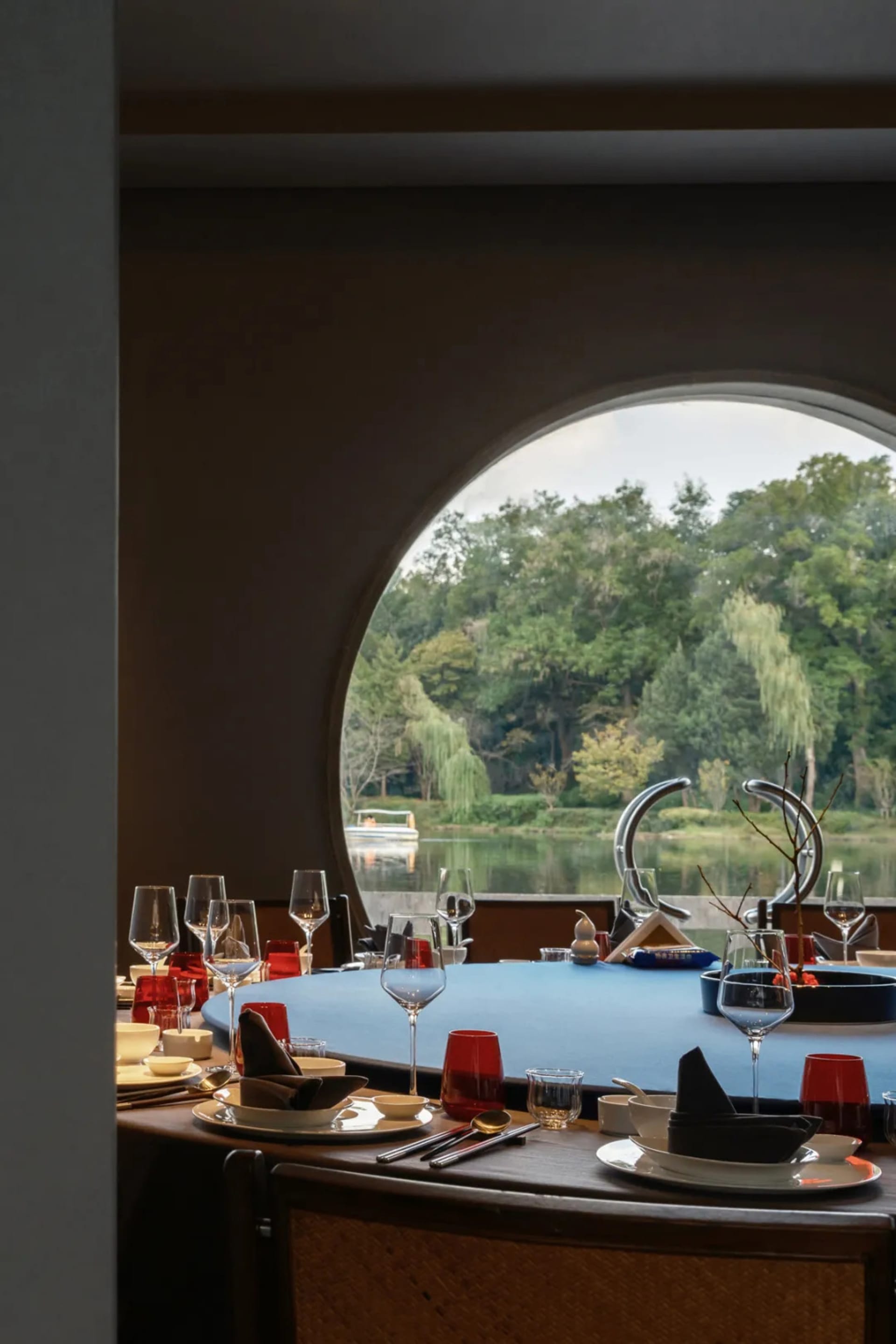 A round table with blue tablecloth and modern decor including wine glasses, plates, and cutlery, set against a scenic lake vi